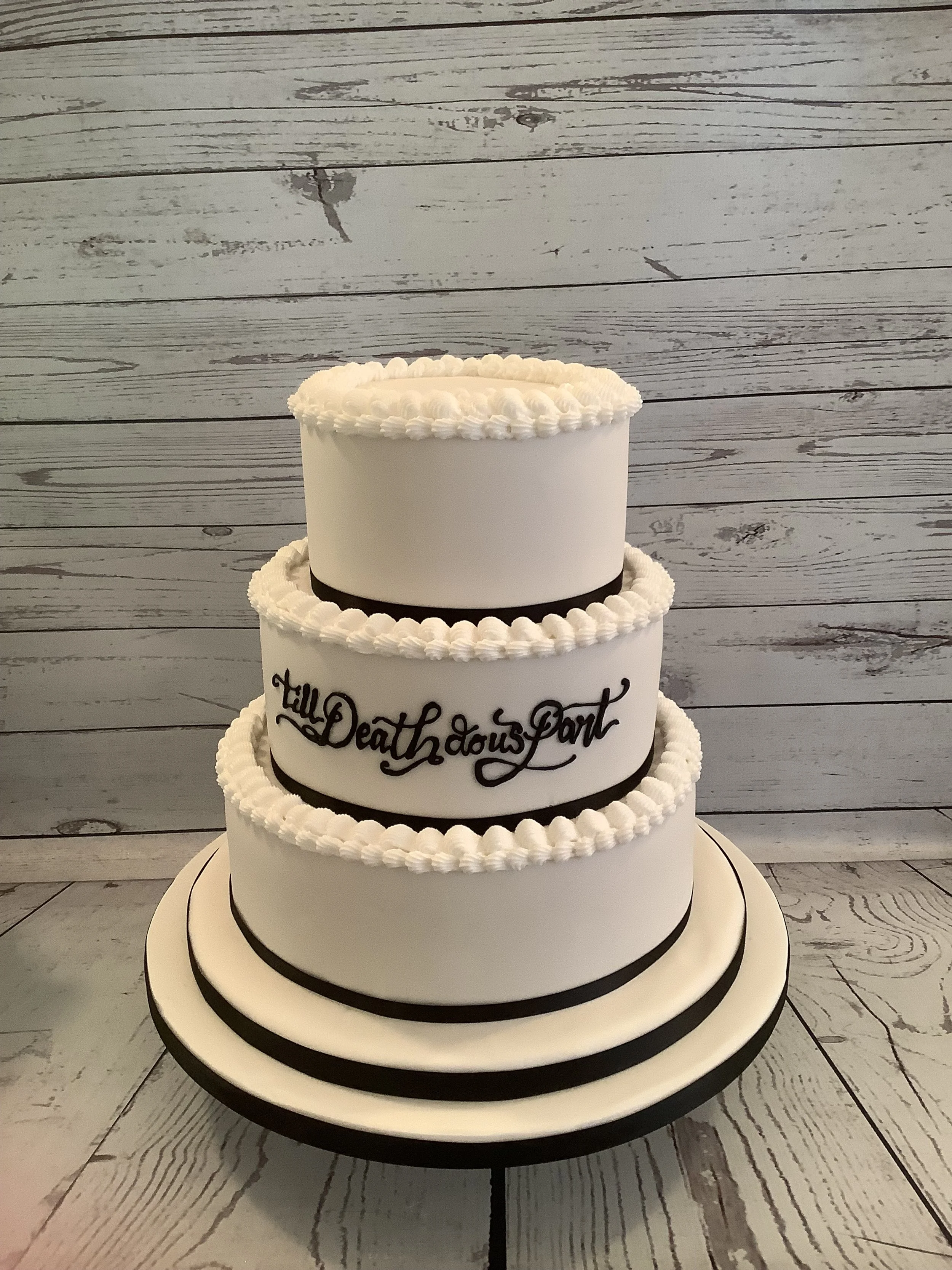 A three-tiered white wedding cake with black accents and decorations, including black ribbons and piping, placed on a white plate with black trim, and set against a wooden background.