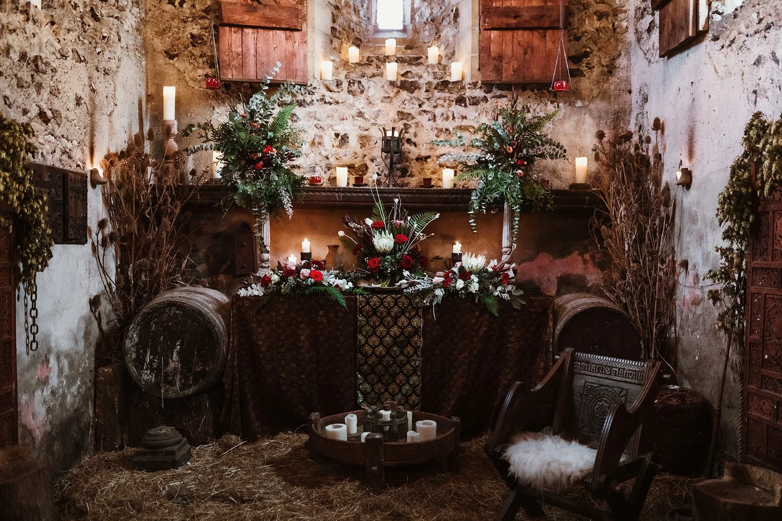 Rustic altar decorated with candles and floral arrangements, set in a stone-walled room with hay-covered floor, featuring barrels, wooden chairs, and dried branches.