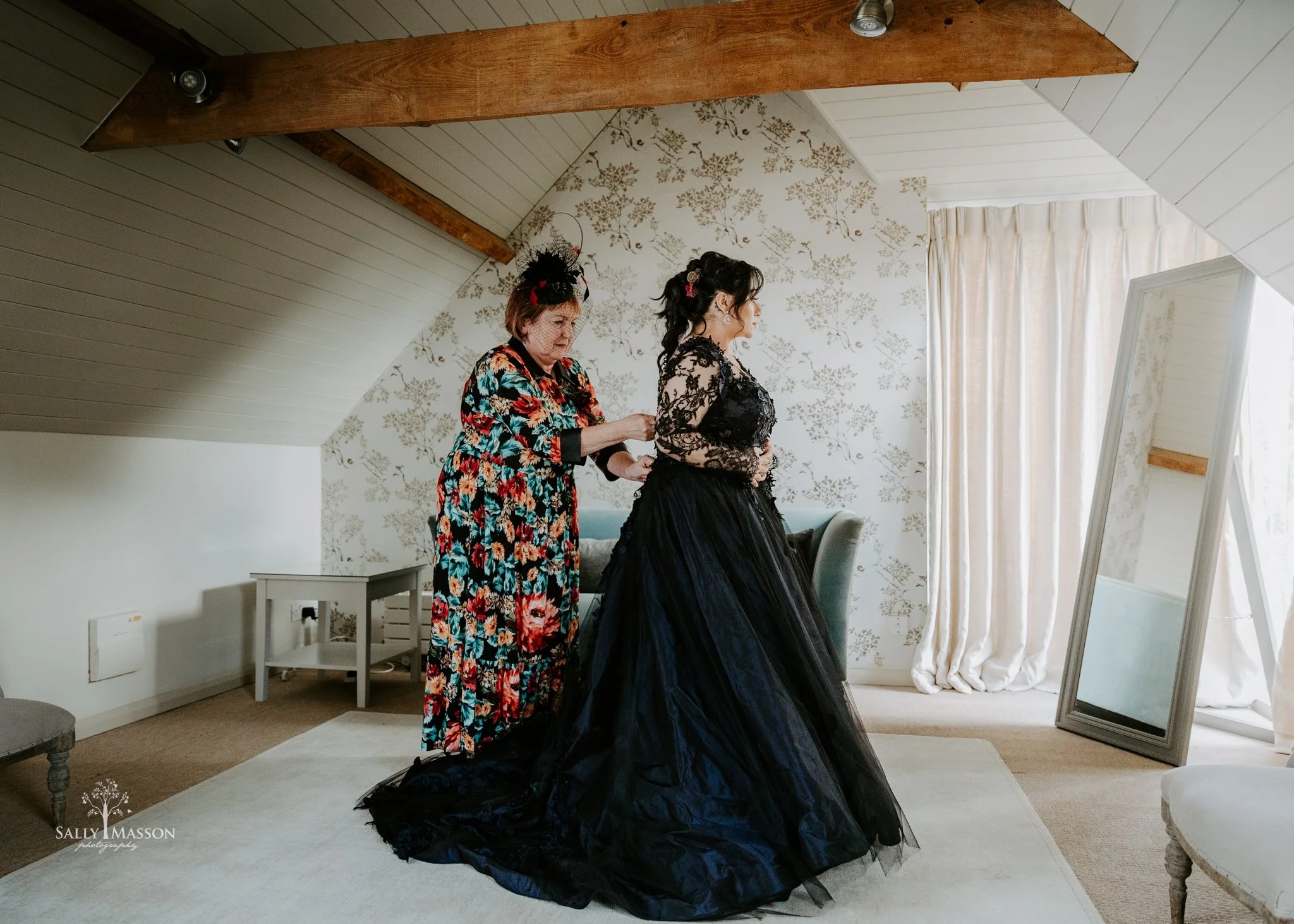 A woman in a black lace gown standing in front of a mirror, while another woman in a floral dress helps her with her dress in a room with floral wallpaper, curtains, and a full-length mirror.