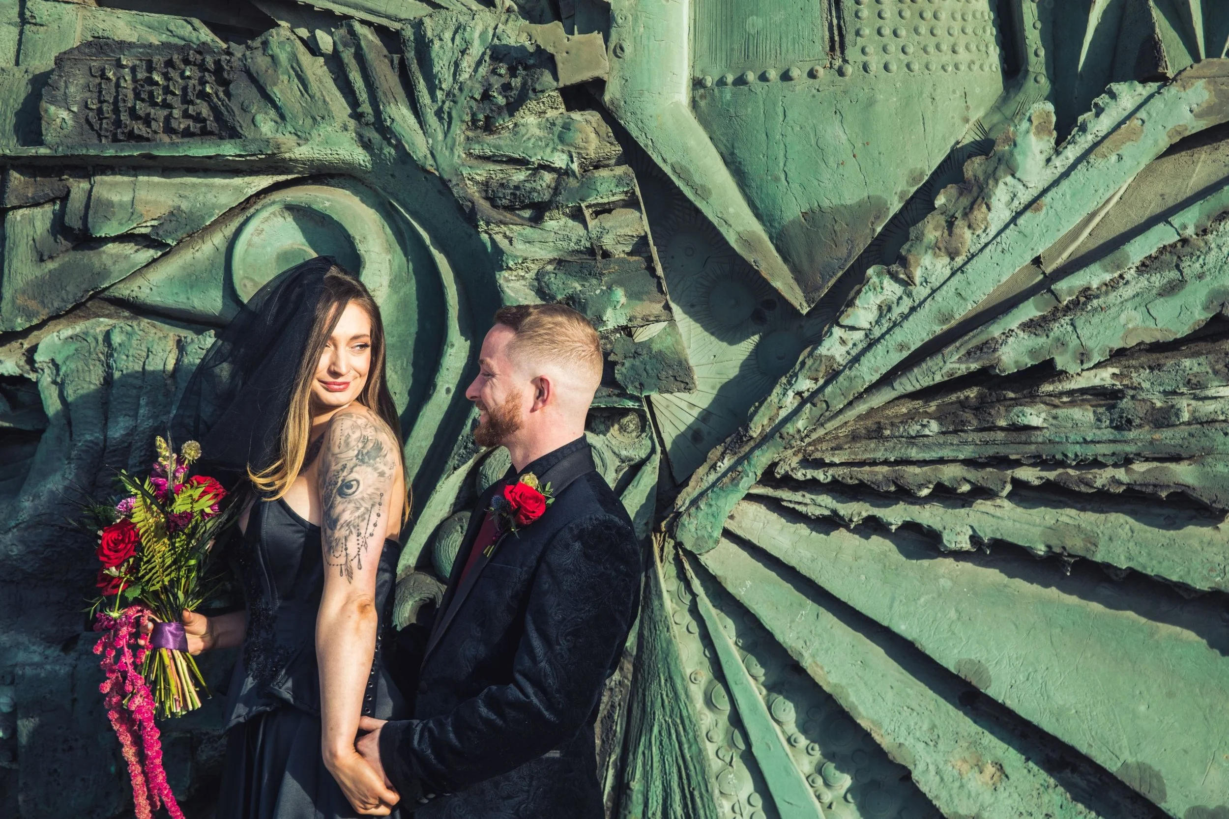 A couple stands in front of a large, textured, metallic green sculpture. The woman has long hair, tattoos on her arm, and is holding a bouquet of flowers. The man has short hair, a beard, and is wearing a suit with a red rose boutonniere. They are ho