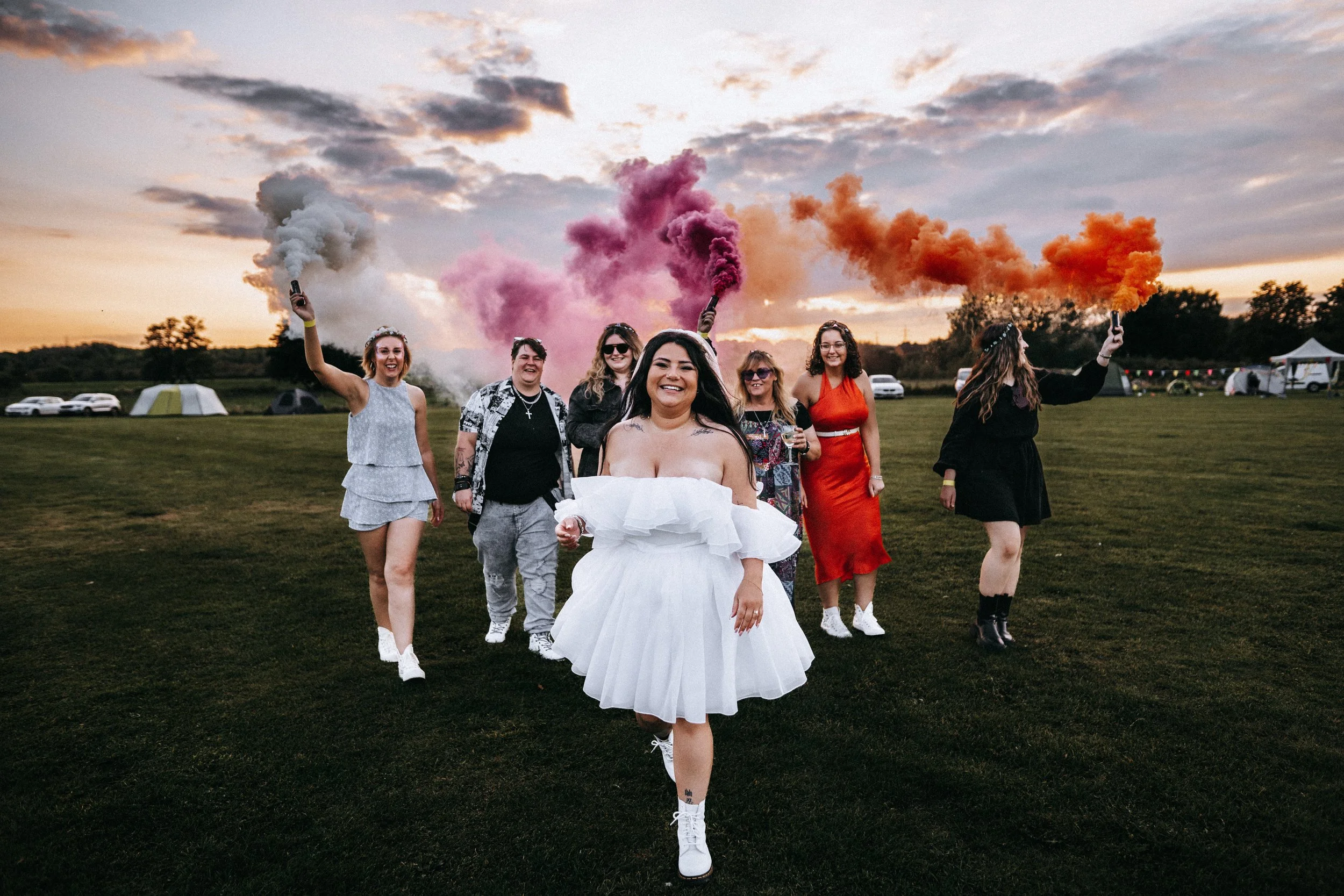A group of women walking outdoors at sunset, with some holding colored smoke flares, celebrating.