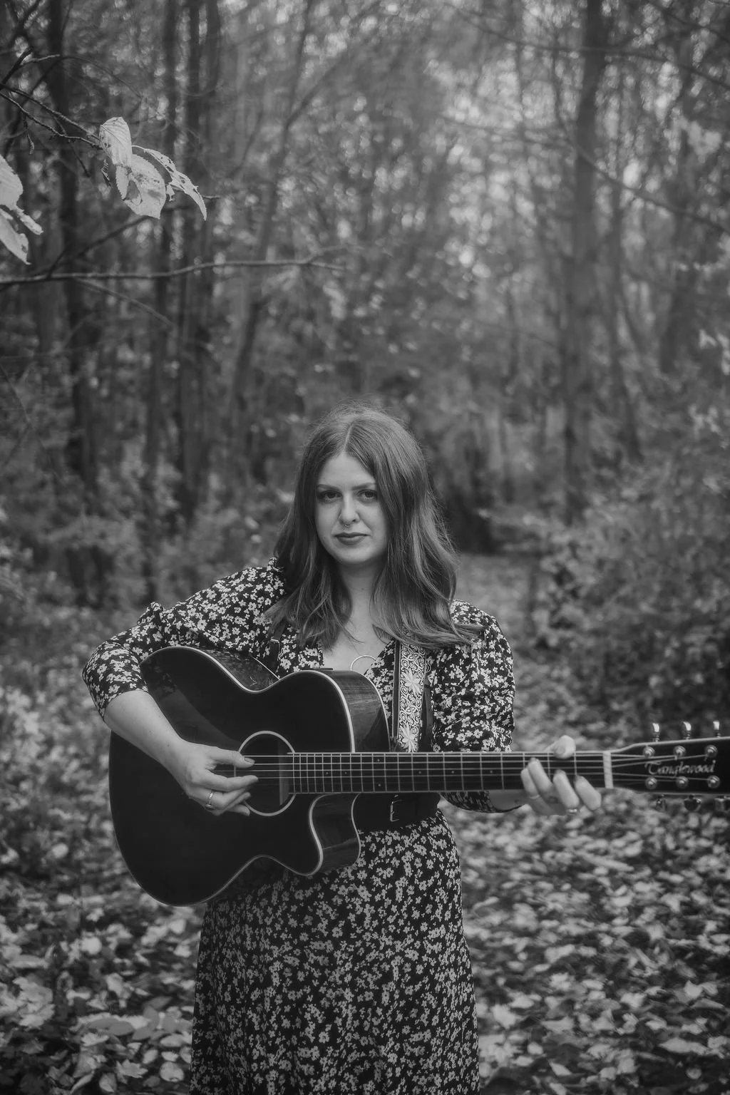 A woman with wavy hair wearing a floral dress playing an acoustic guitar outdoors in a wooded area.