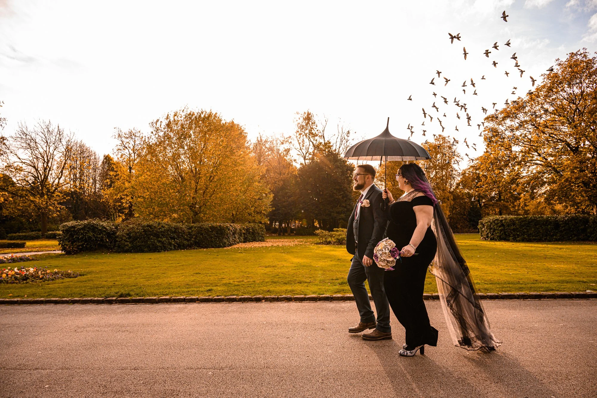 A couple dressed in formal attire walking in a park during autumn, with a woman holding a bouquet of flowers and an umbrella, and birds flying in the sky.