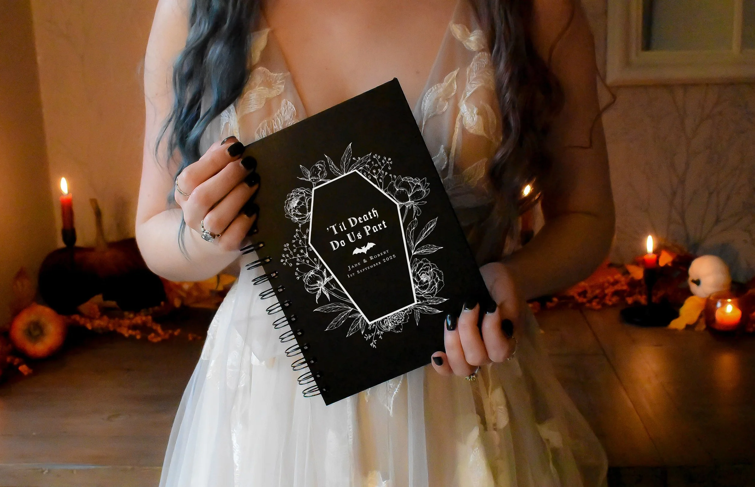 Person holding a black notebook with white floral design and text 'Til Death Do Us Part' at a Halloween-themed setup with candles and pumpkins in the background.