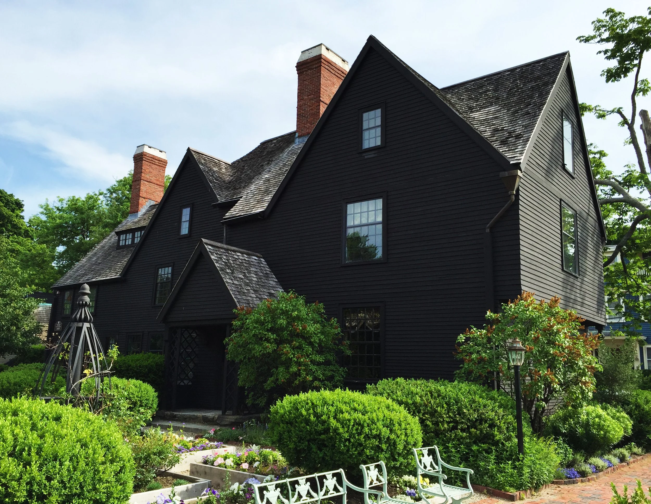 Black historic house with steep gabled roof, brick chimneys, surrounded by lush green bushes, trees, and a garden with flower beds, white benches, and a decorative garden structure.