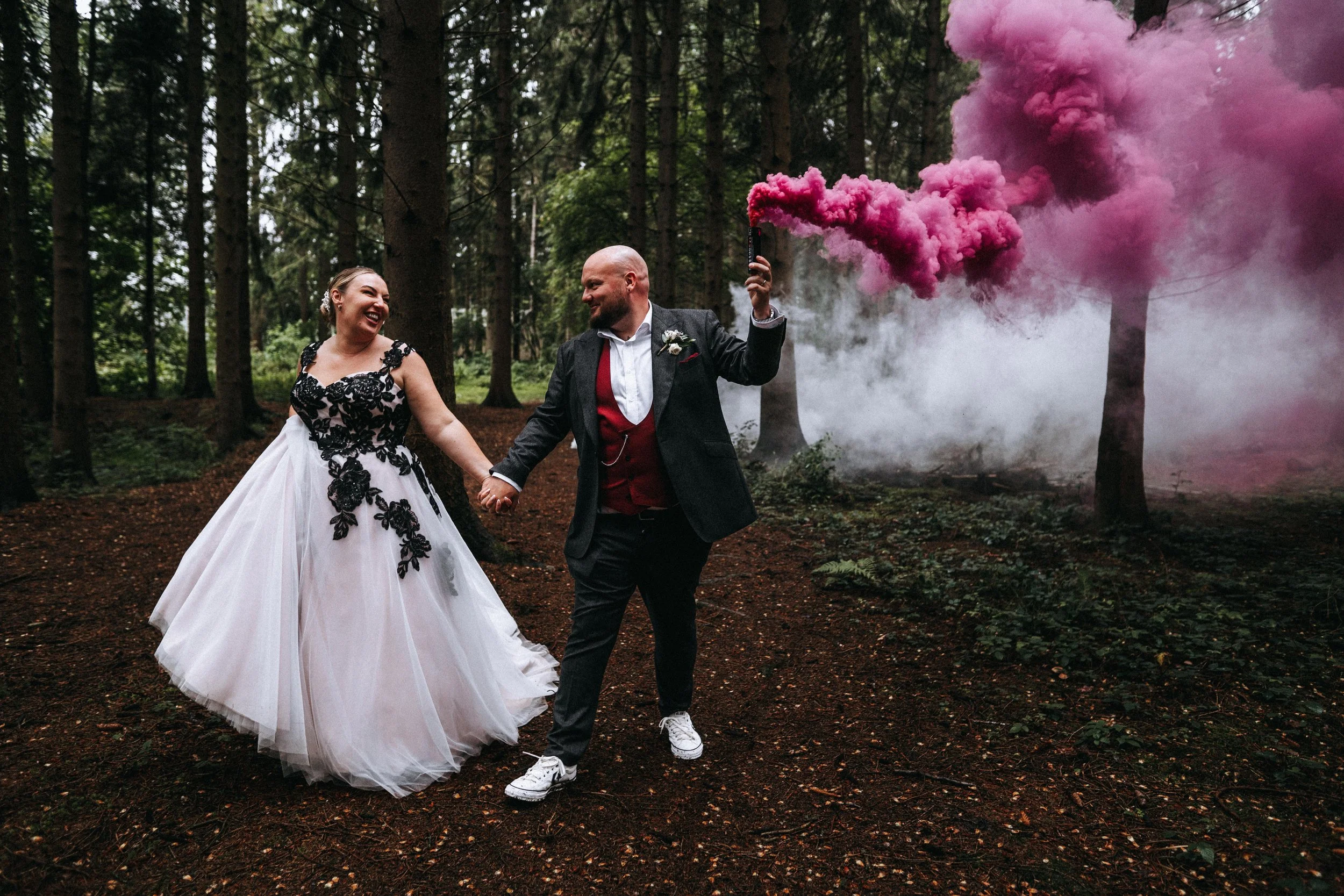 A newlywed couple, smiling and holding hands, walking through a forest with pink smoke in the background.