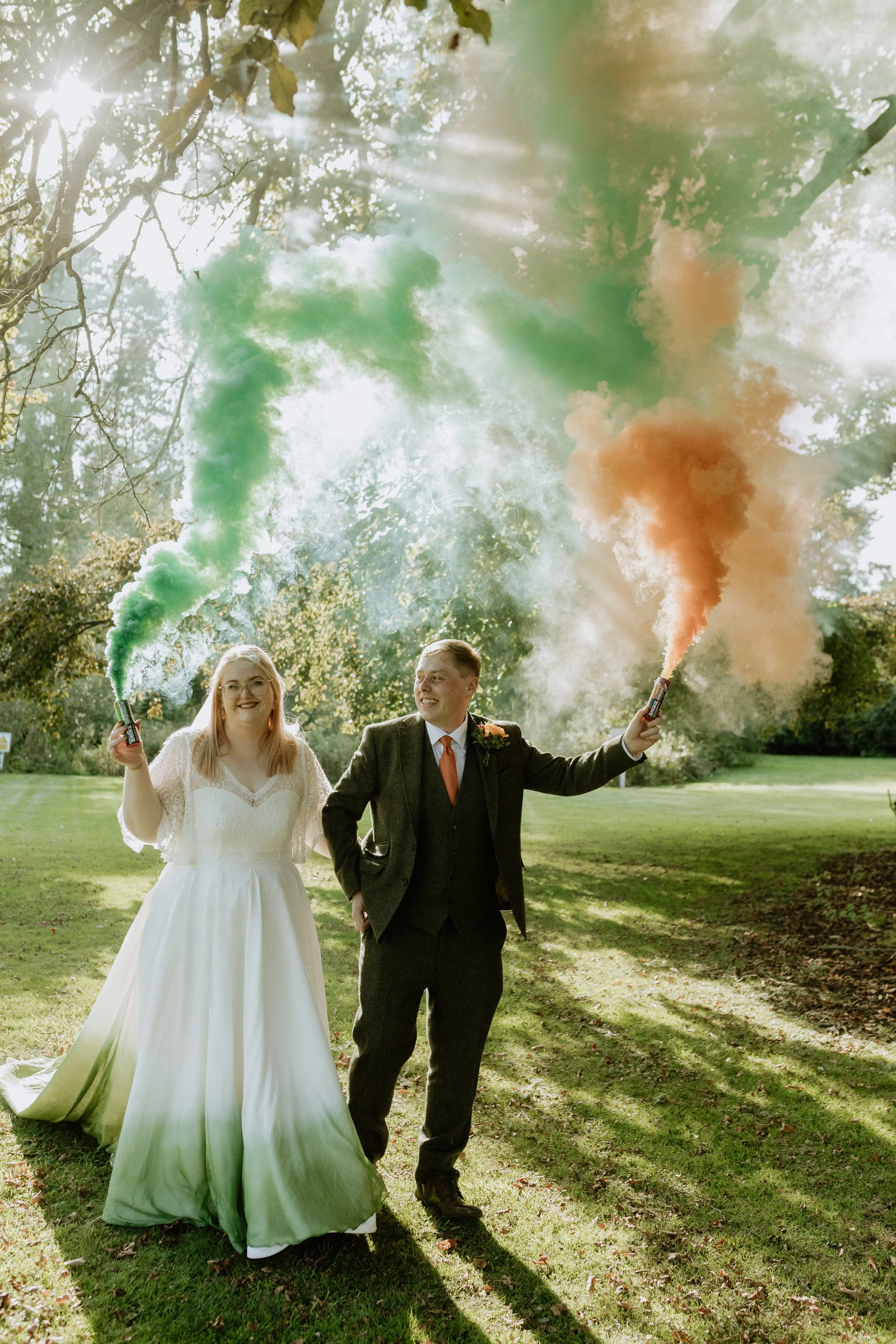 A smiling couple walking outdoors in a park, celebrating their wedding with colorful smoke bombs in green and orange, with sunlight filtering through trees.