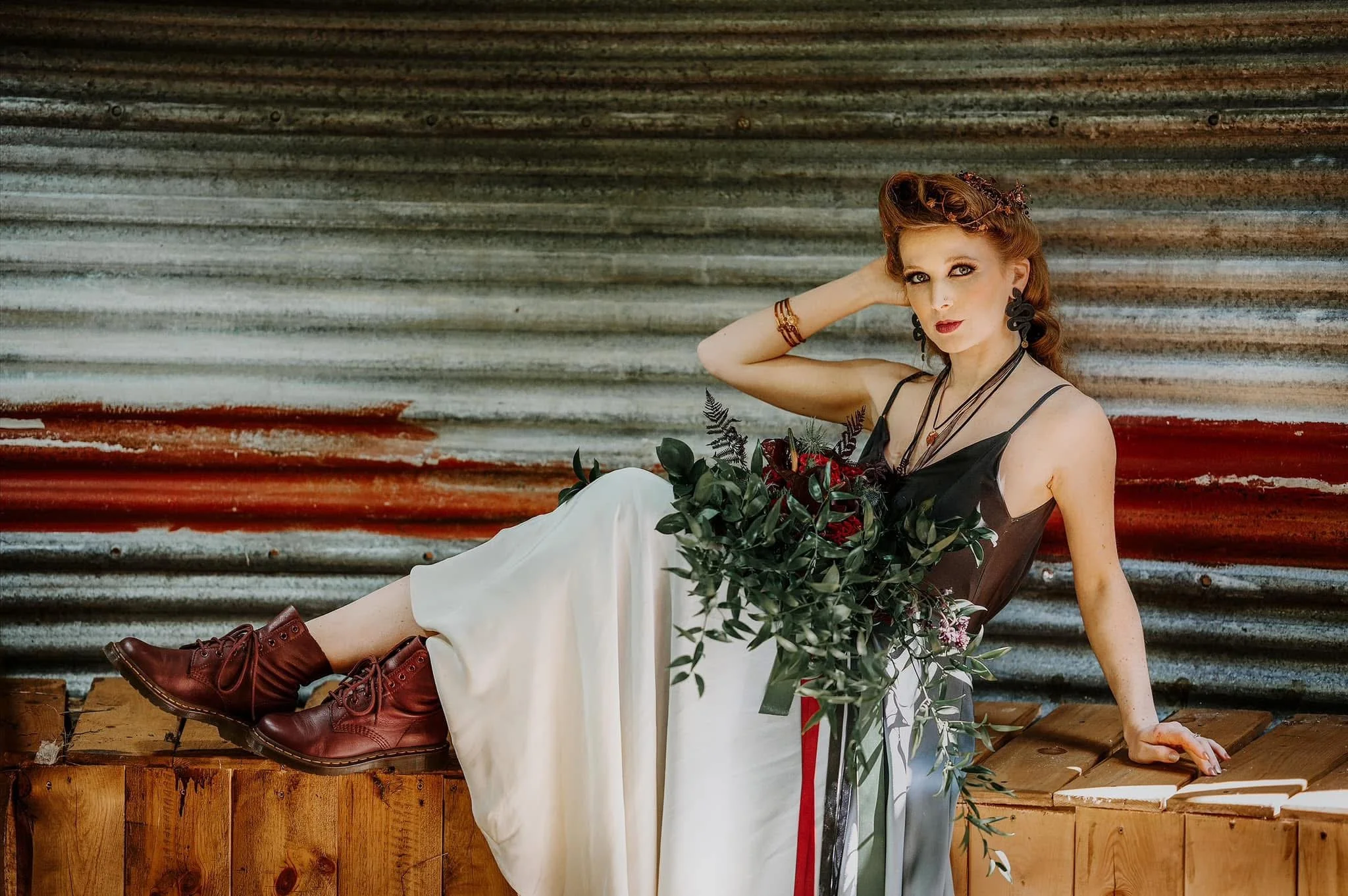 A woman with red hair sitting on a wooden surface against a corrugated metal background. She is wearing a black spaghetti strap top, white pants, red boots, and holding a bouquet of red roses and greenery. She has jewelry, including earrings and brac