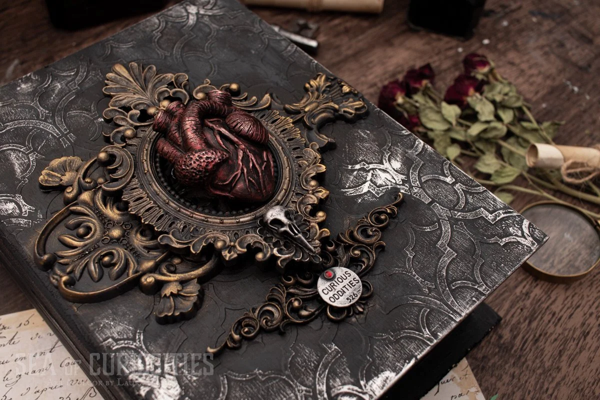 Decorative ornate box with a detailed anatomical heart sculpture on top, surrounded by intricate gold-colored scrollwork, alongside a skull and a tag labeled 'Curious Oddities 526'. In the background, dried roses and a magnifying glass are on a wooden surface.