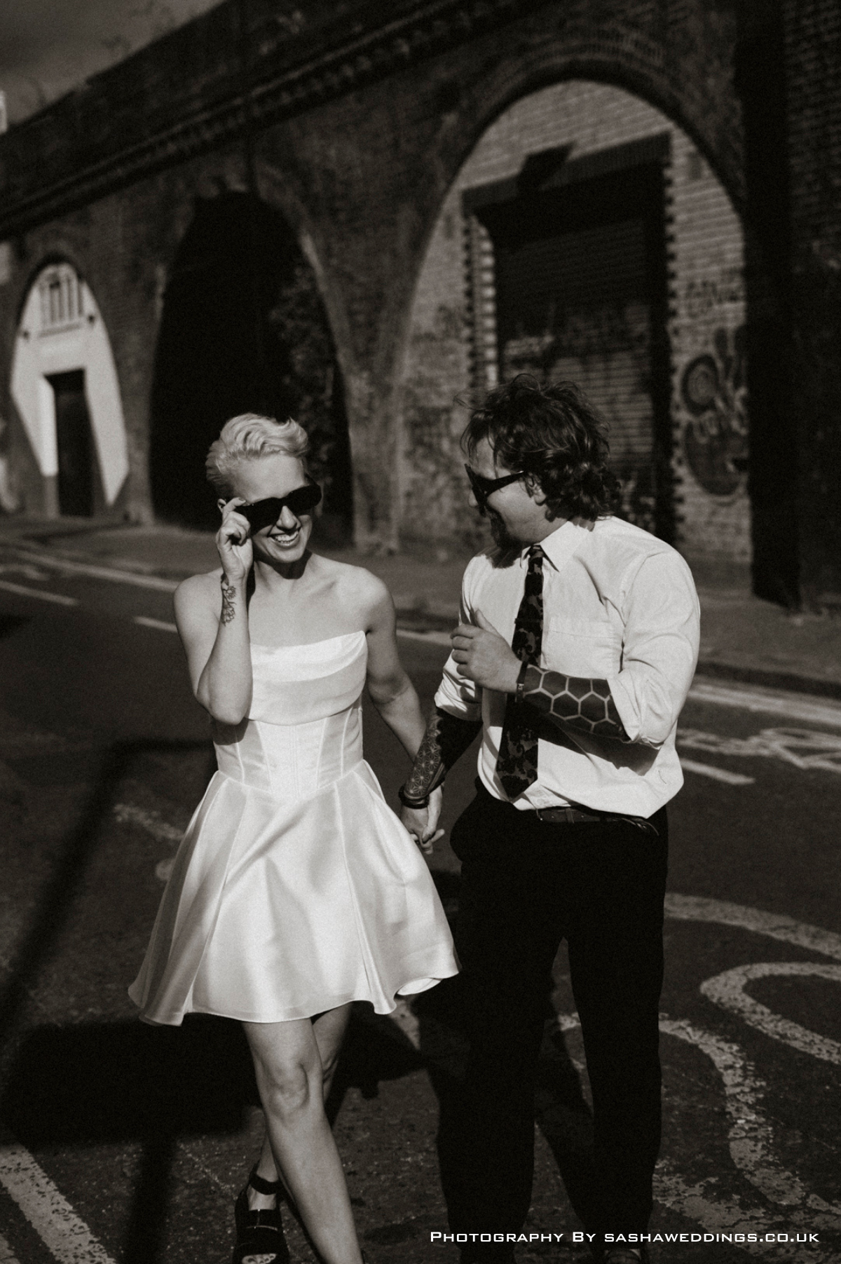 A black and white photo of a smiling woman in a strapless dress and sunglasses, holding hands with a man in a shirt and tie, walking together on a city street at night, with a brick archway background.