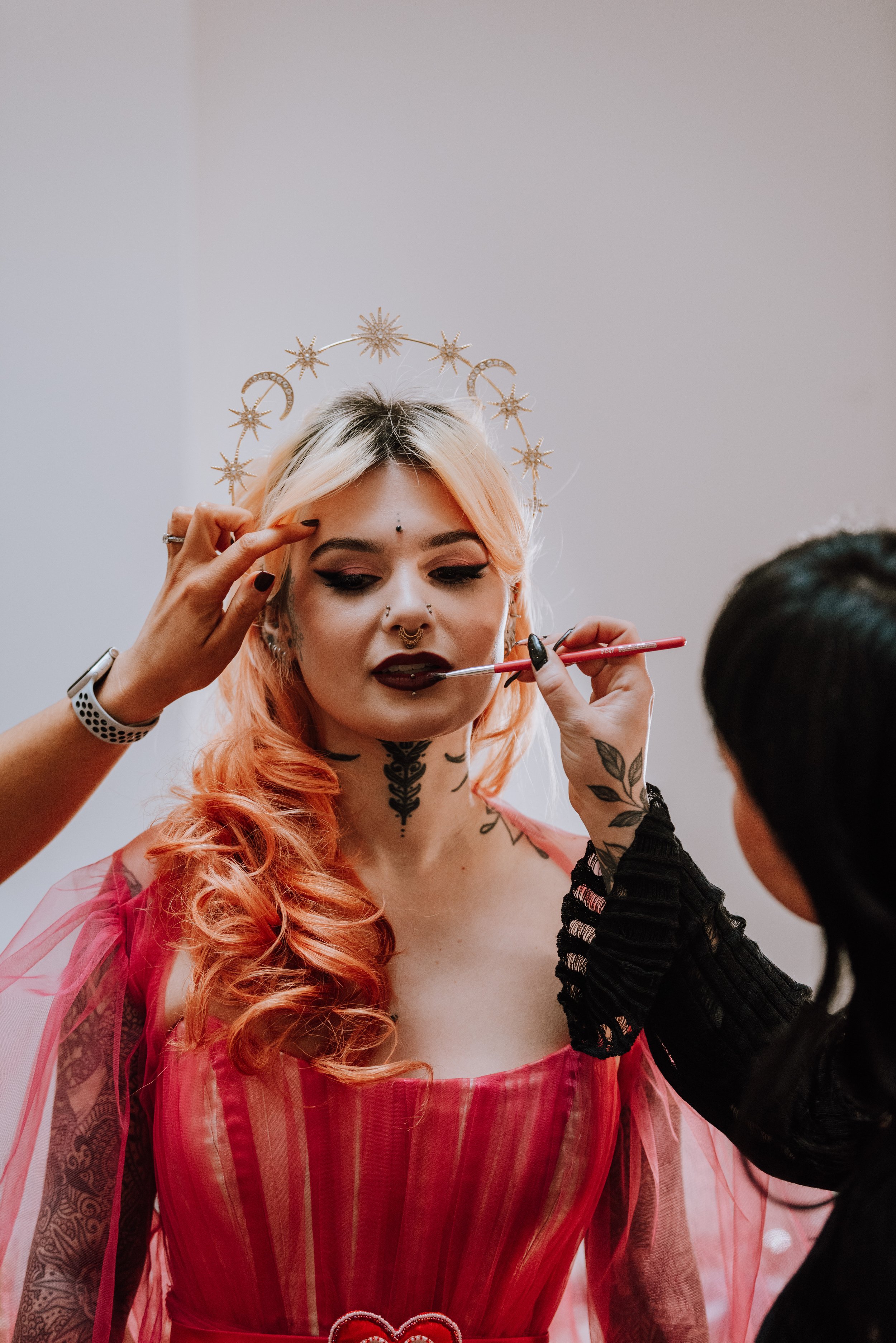 A woman with pink hair, tattoos, and gothic makeup is having her lips painted by a makeup artist. She is wearing an ornate headpiece and a red dress with mesh sleeves, standing against a plain background.
