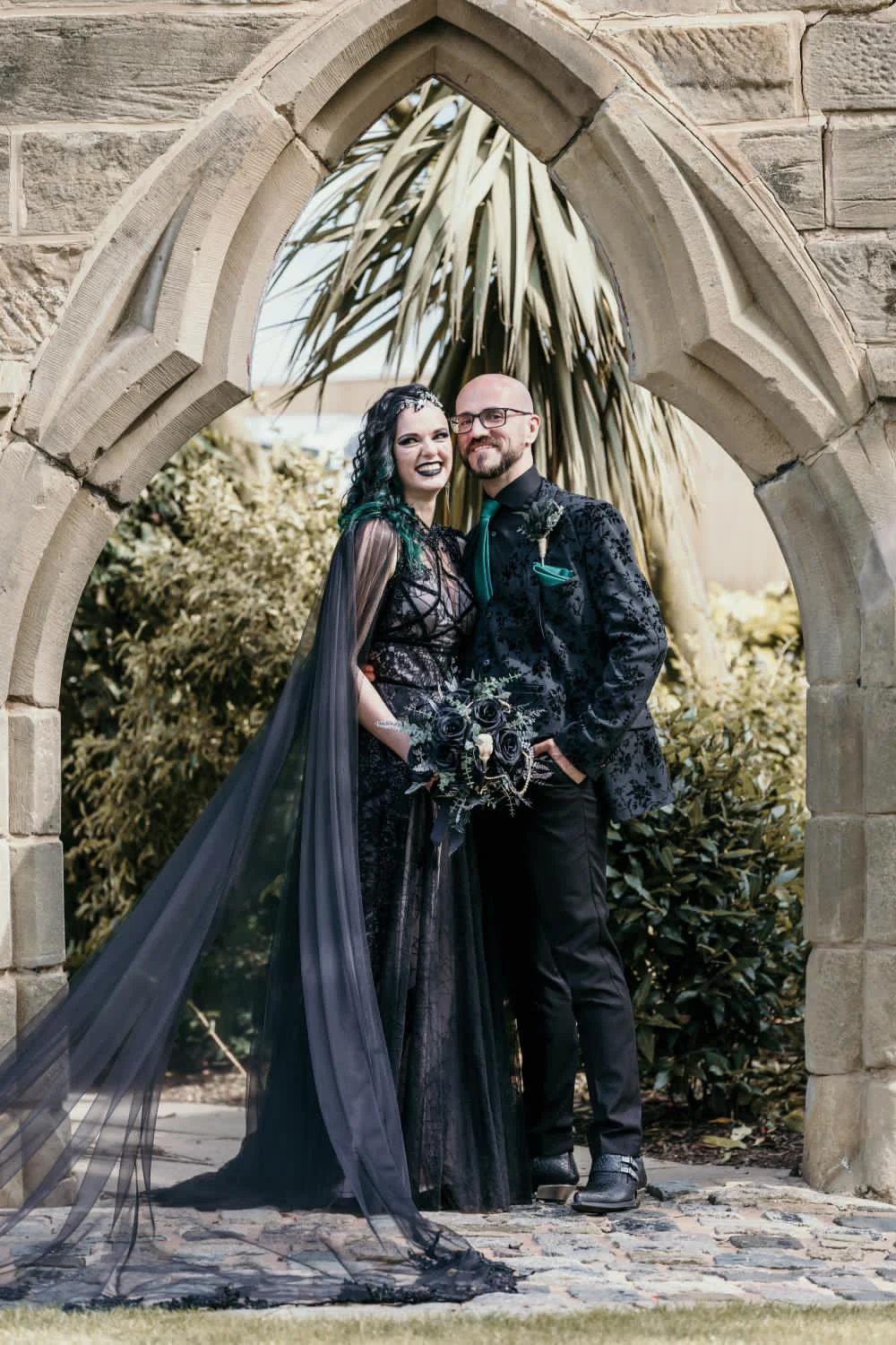 A gothic couple dressed in black standing under an arched stone structure with plants in the background.