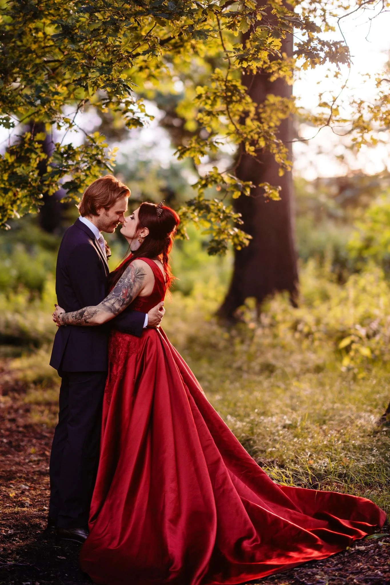 A couple dressed in formal attire embracing each other outdoors during sunset, with green trees and sunlight in the background.