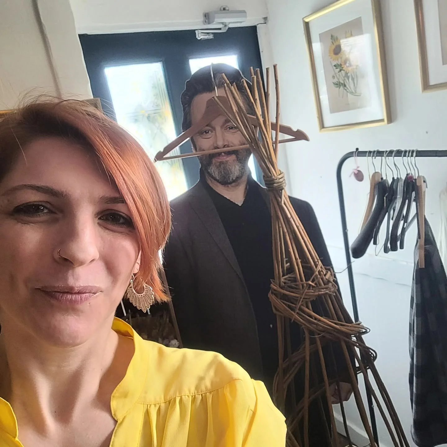 A woman with reddish hair and wearing a yellow top takes a selfie indoors, with a man behind her dressed in a suit, holding a large bundle of wicker sticks and a wooden hanger with more wicker sticks. The background includes a clothing rack with vari