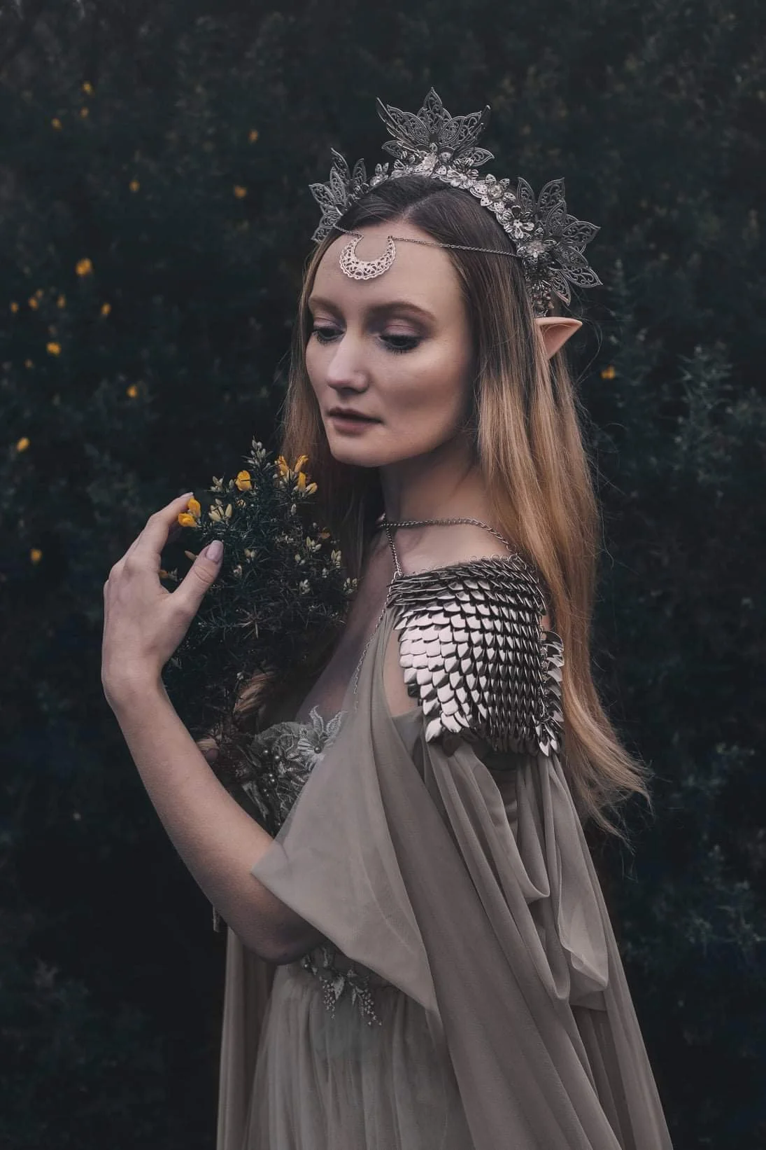 A woman with elf-like ears, wearing a silver crown and jewelry, dressed in a flowing beige gown with metallic shoulder accents, holding a small bouquet of yellow flowers, standing outdoors at dusk.