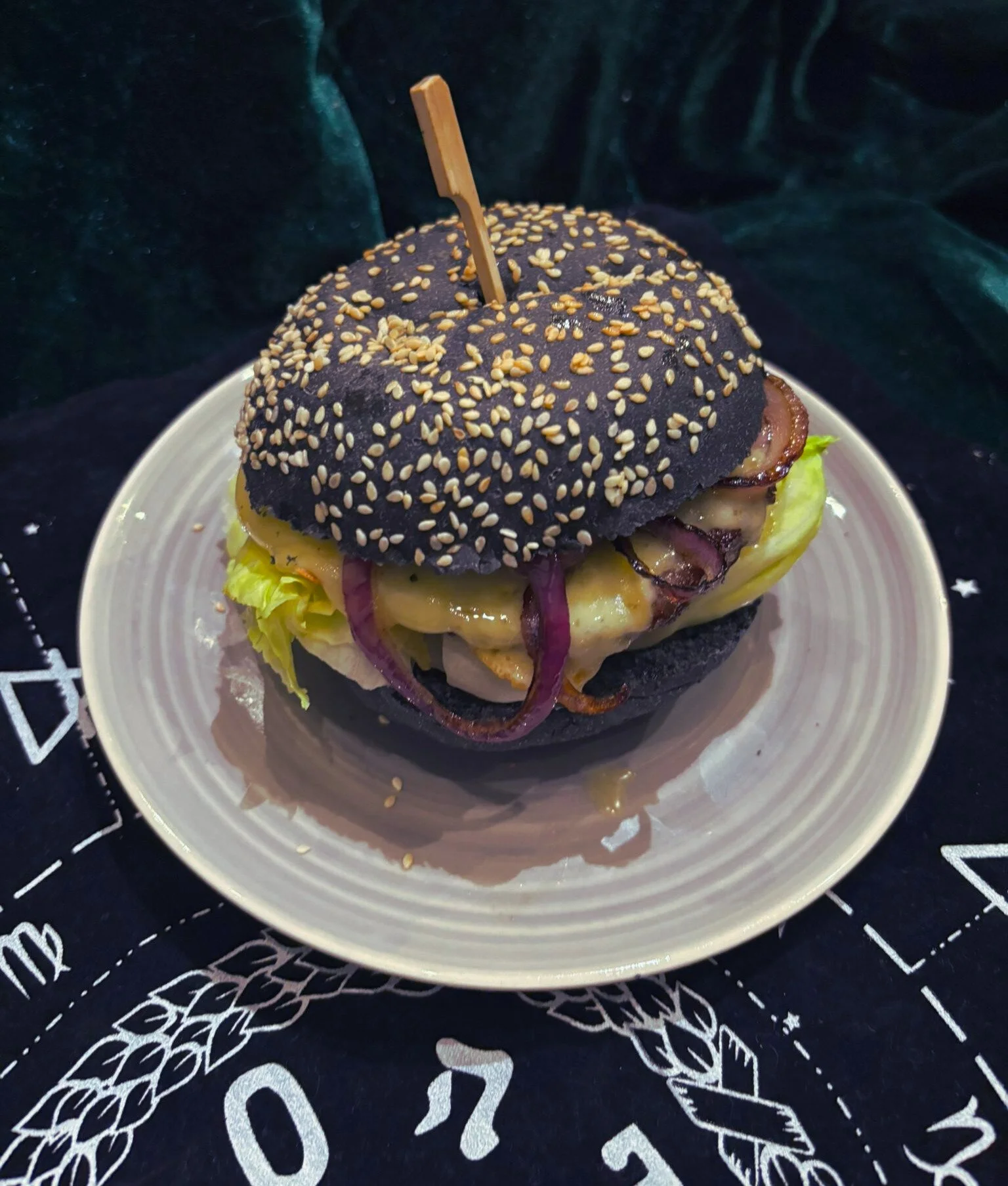 Black burger bun with sesame seeds, containing lettuce, cheese, bacon, and red onions, served on a white plate.