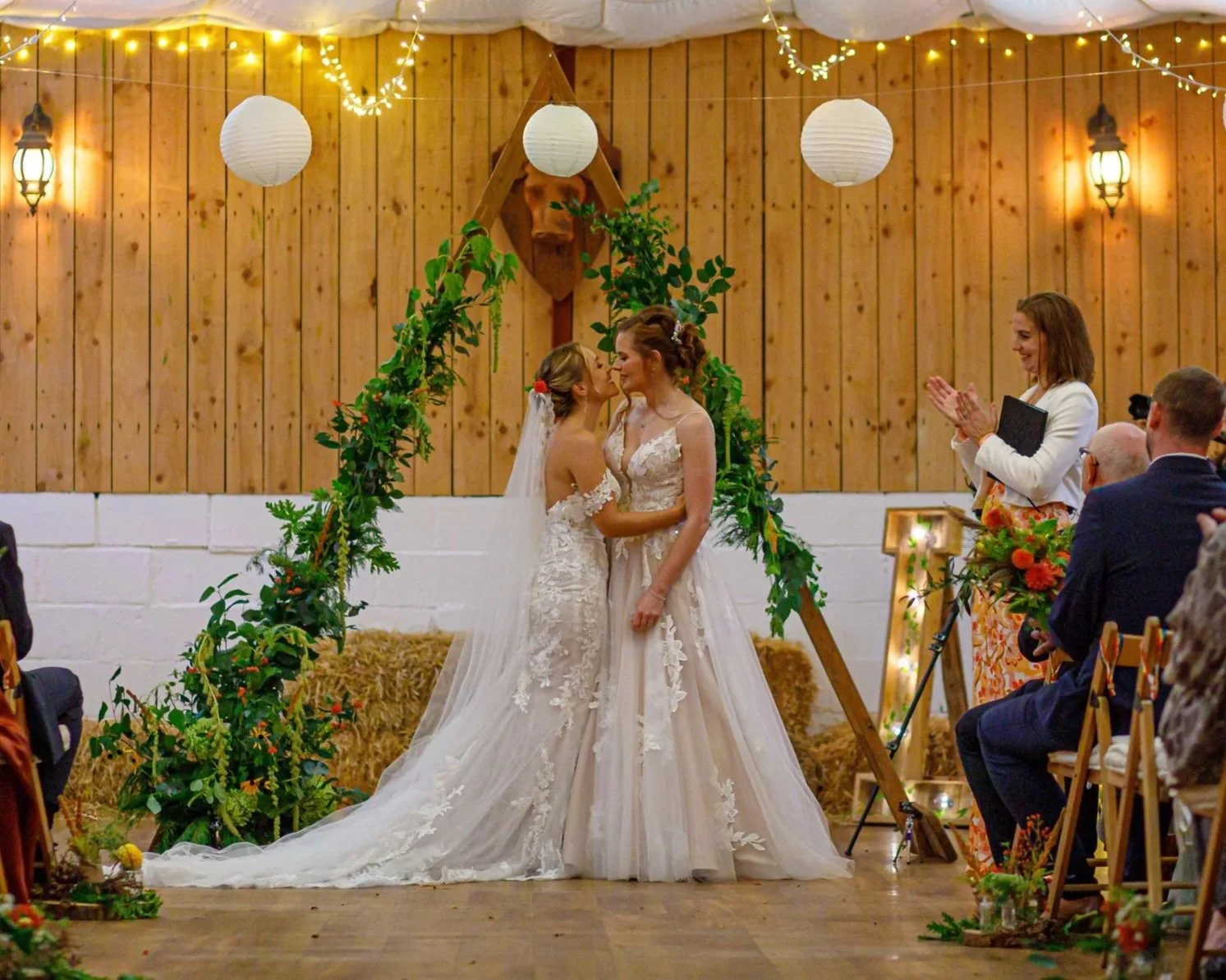 Two brides in wedding dresses standing together under a floral arch, holding hands, during a wedding ceremony in a rustic indoor venue with wooden walls and hanging lanterns. Guests are seated on either side, and a woman claps on the right.
