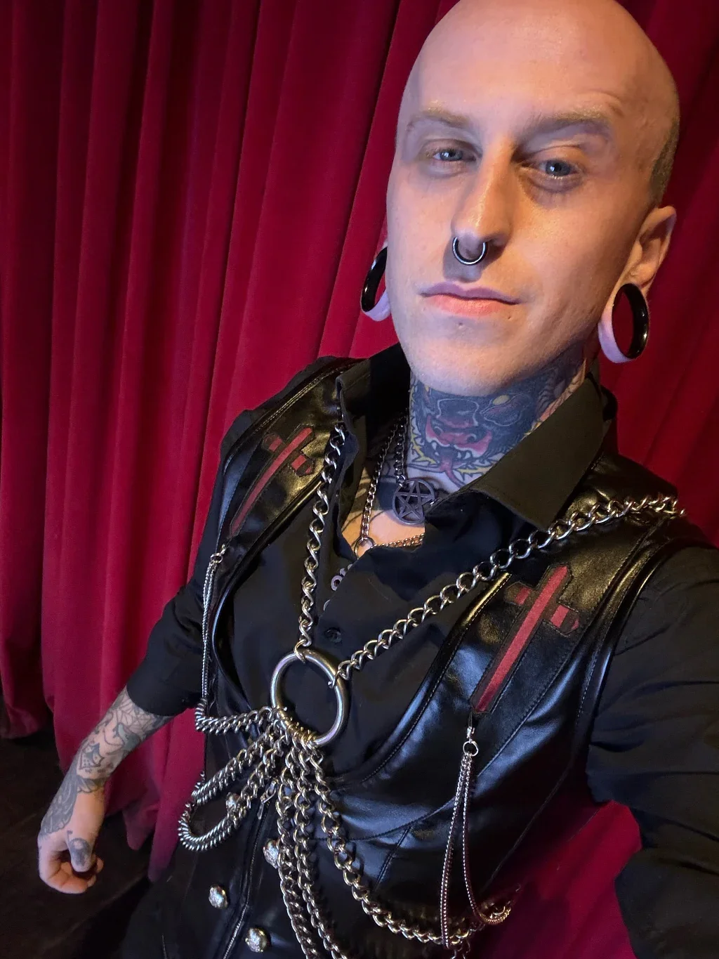 A person with a bald head, multiple facial piercings, neck tattoos, and wearing black clothing with chains and accessories, standing in front of red curtains.
