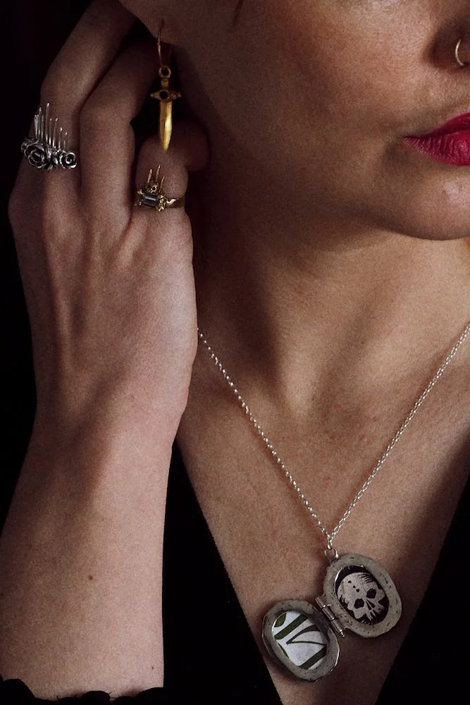 Close-up of a woman wearing multiple rings, gold earrings, a necklace with a locket featuring a skull design, and a septum piercing.