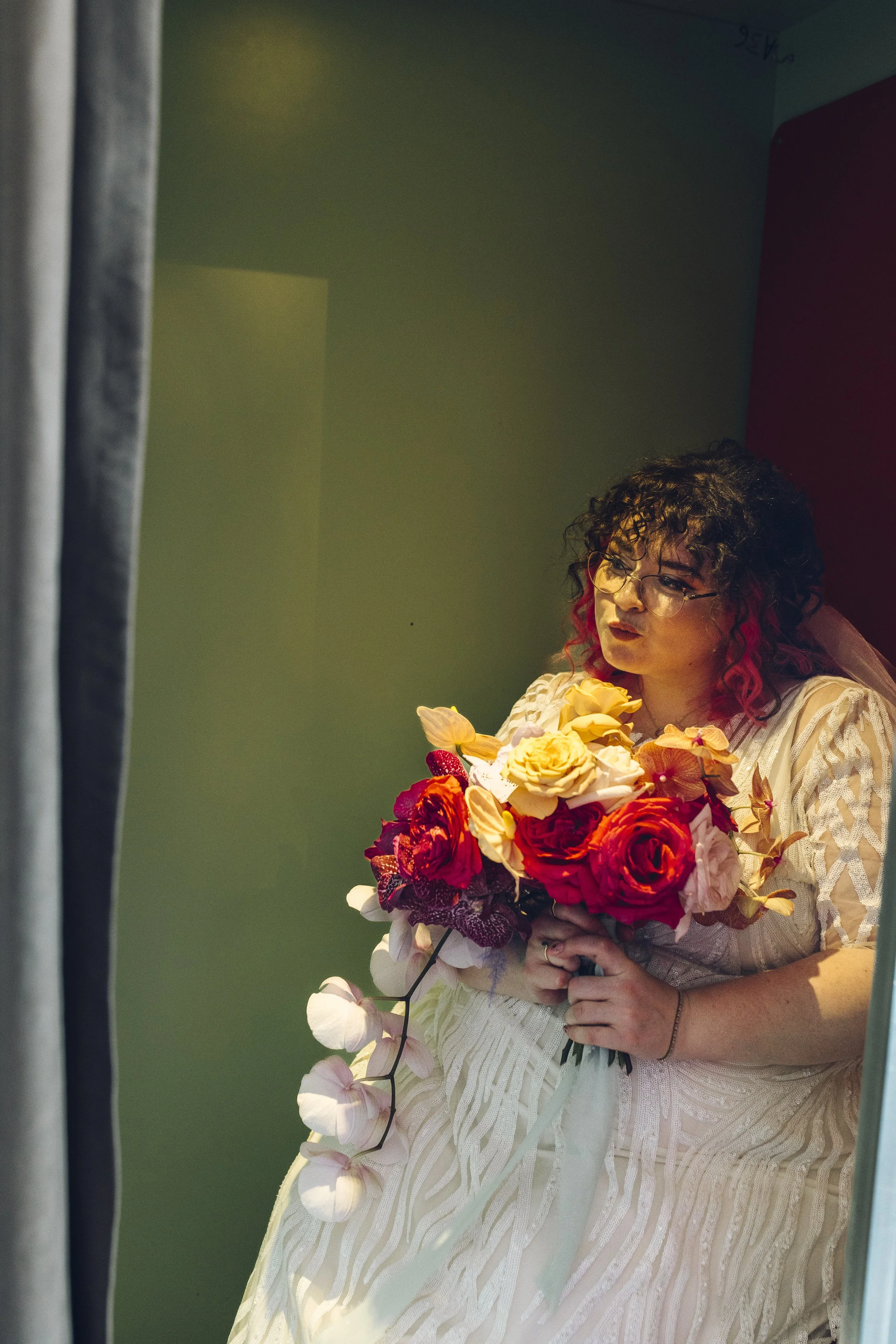 A woman with curly, dark hair with red tips and glasses, wearing a cream-colored dress, holding a large bouquet of roses and other flowers, standing in front of a mirror.