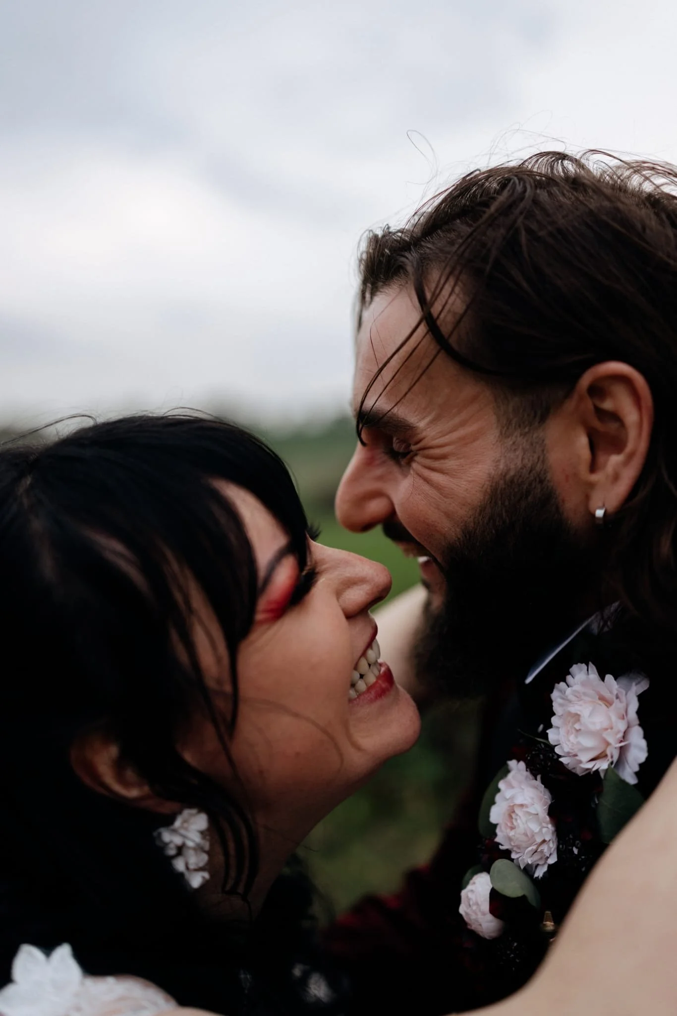 A couple smiling and touching foreheads, outdoors, with cloudy sky in the background.