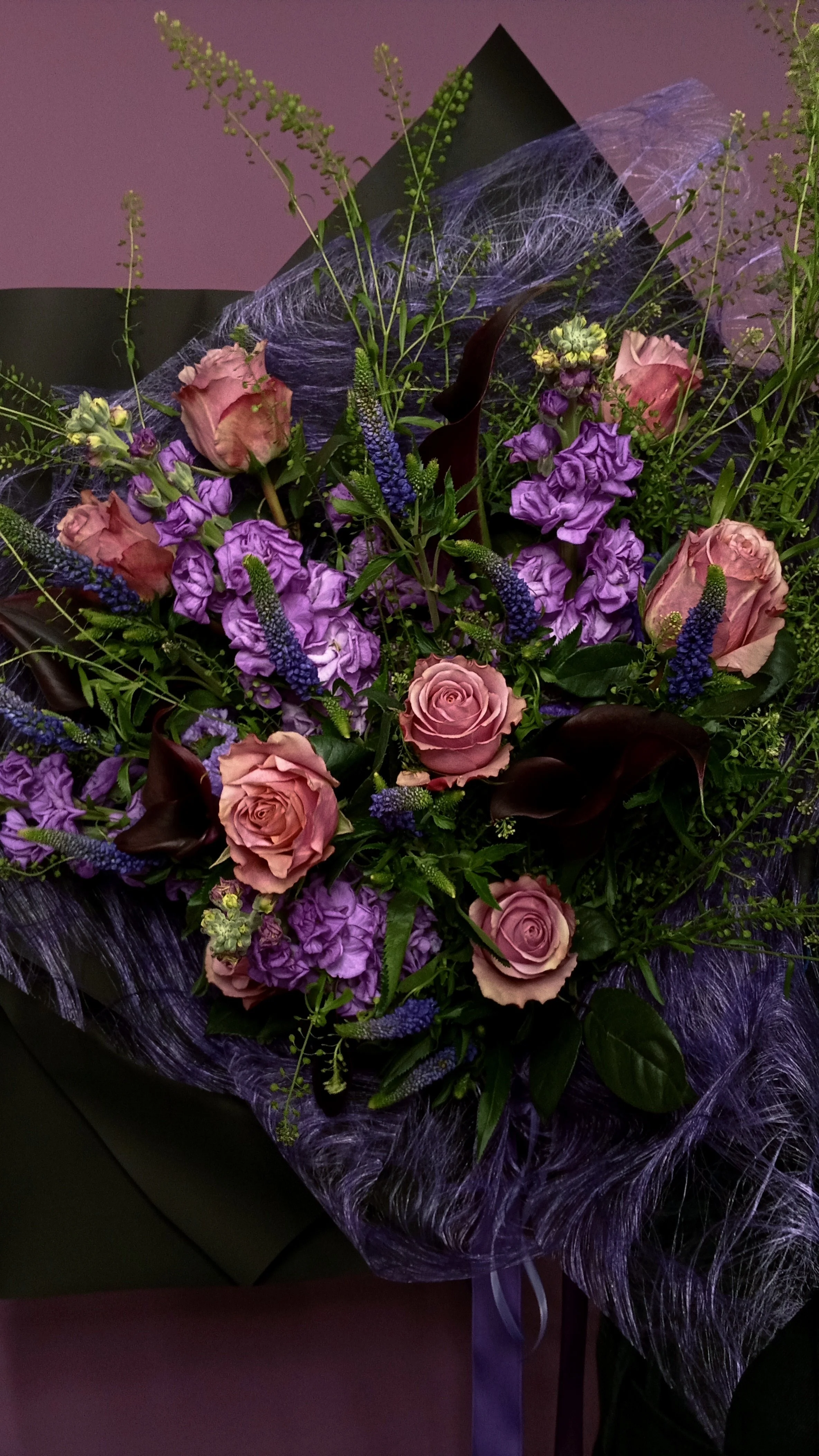A bouquet of pink roses, purple delphiniums, and dark calla lilies wrapped in purple netting.