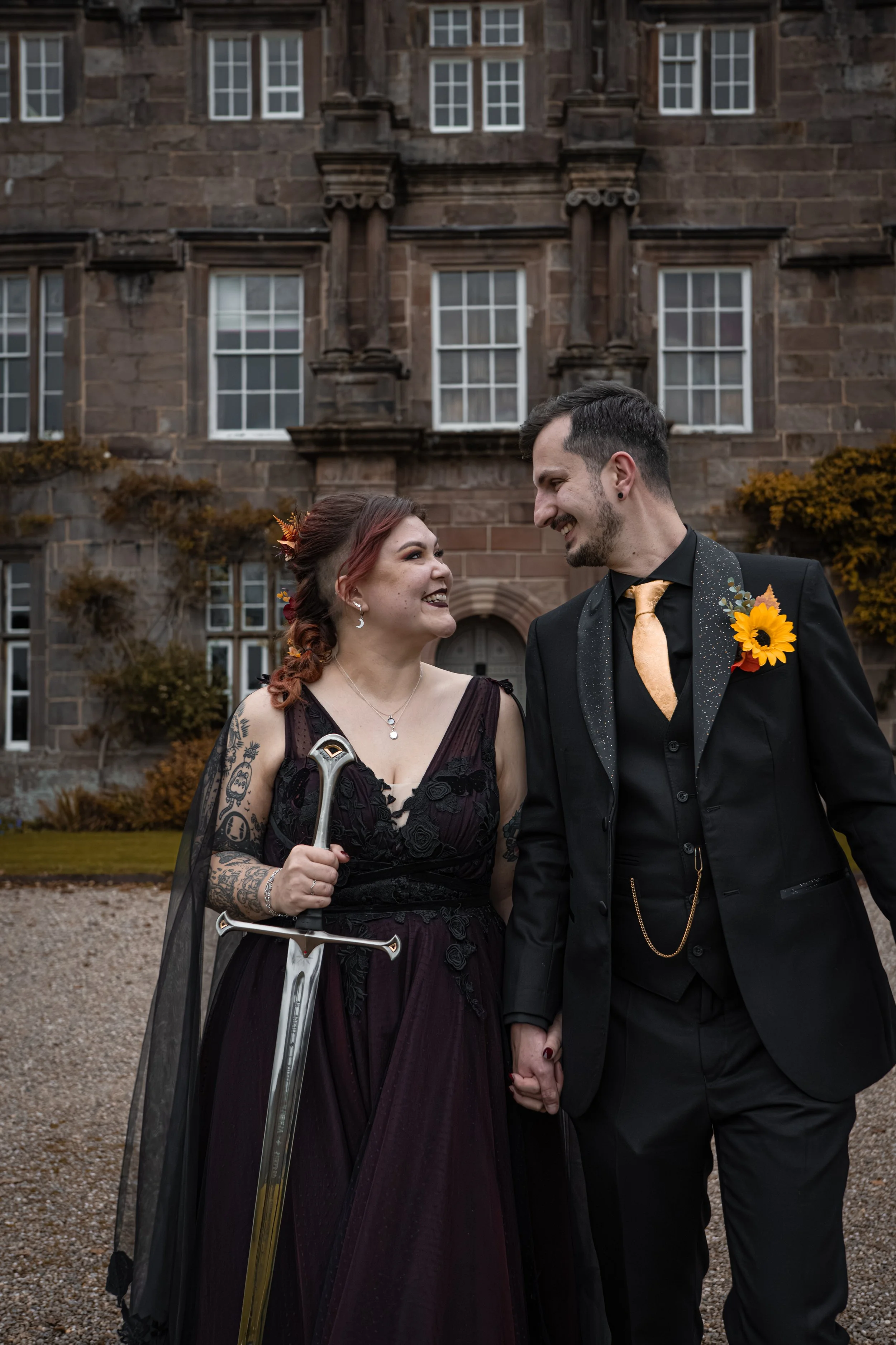 A woman in a dark purple dress and a man in a black suit hold hands and smile at each other outside a large stone building, with the woman holding a sword and wearing a sunflower boutonniere.