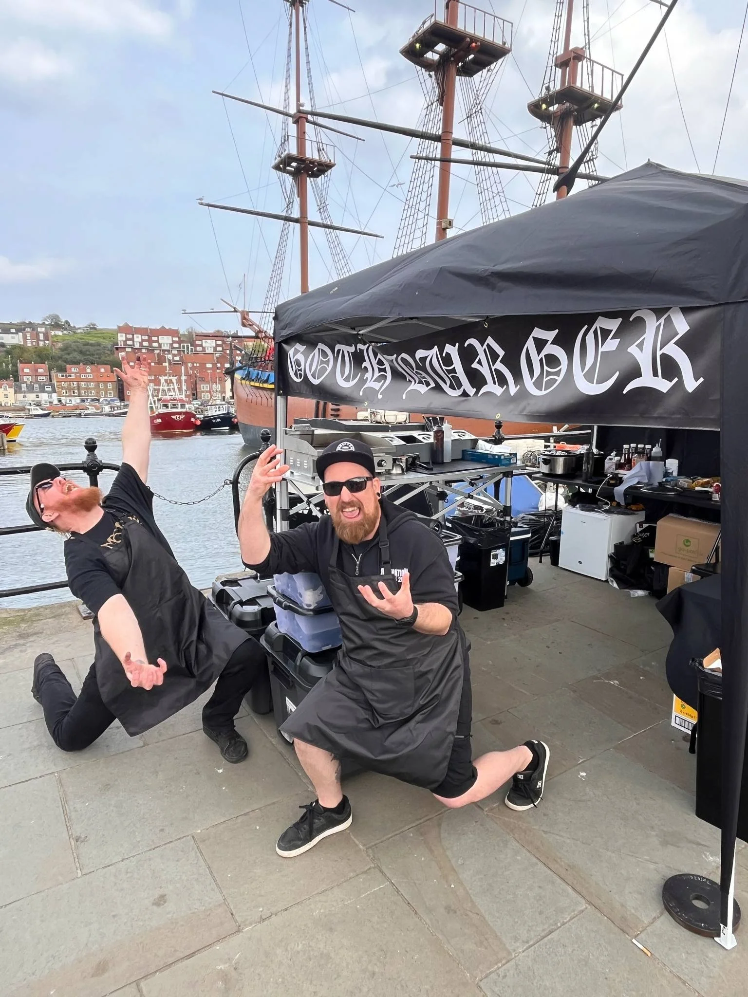 Two men posing in front of a dock and a large historical ship. One man is kneeling and gesturing with one hand, wearing sunglasses and a black apron. The other man is also kneeling with one knee on the ground, wearing sunglasses and a black apron, wi