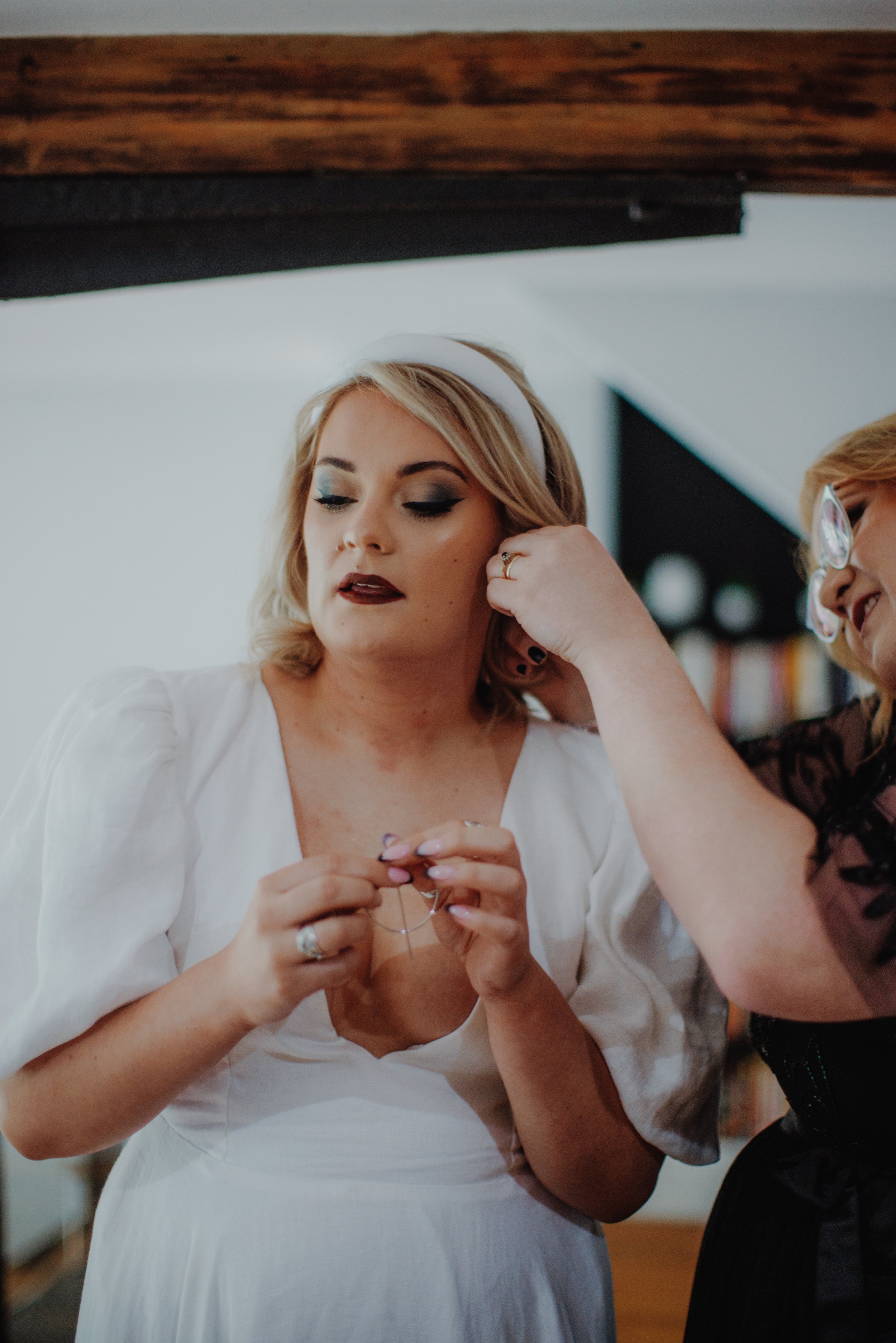 A woman in a white dress is putting on a necklace while another woman adjusts her earrings.