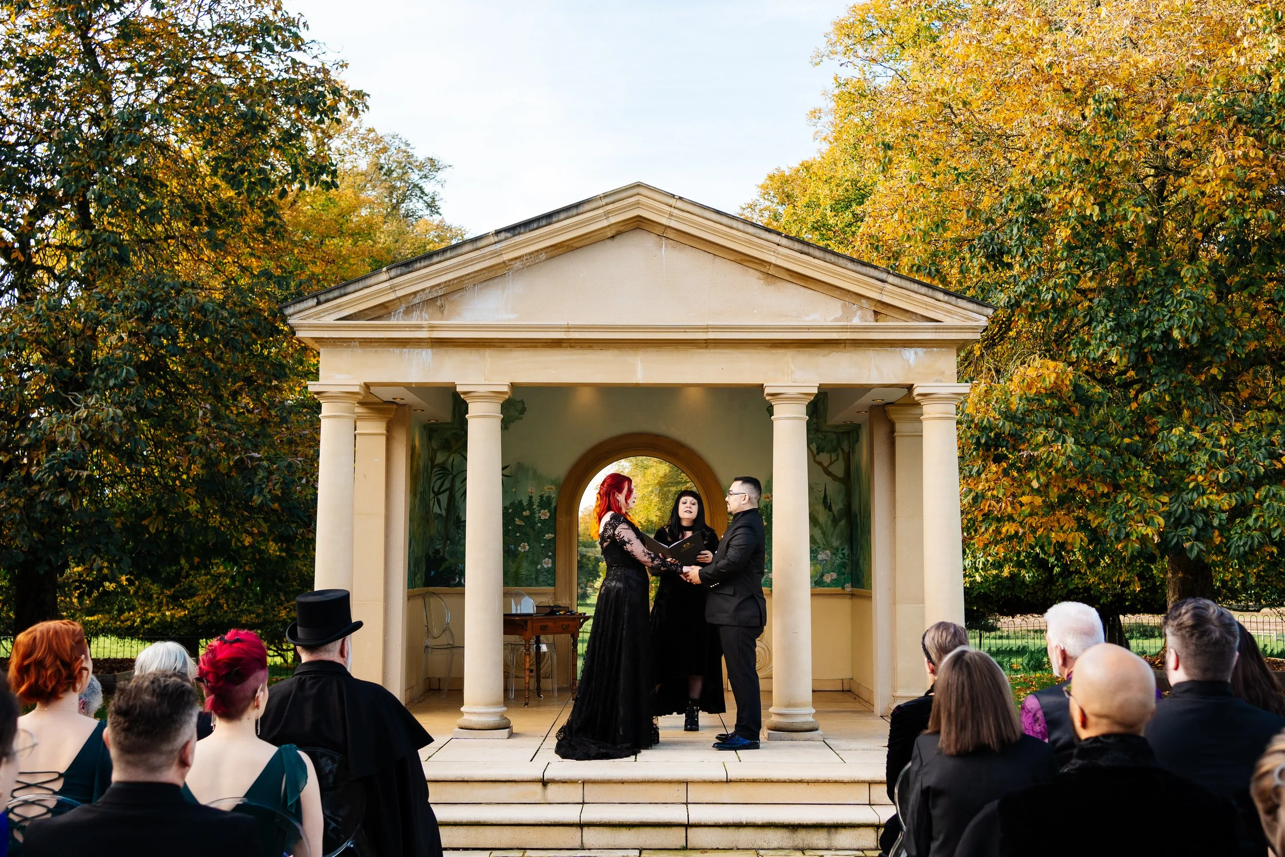 A wedding ceremony taking place outdoors under a small classical pavilion with columns, with a bride and groom standing before an officiant, surrounded by seated guests, autumn trees in the background.