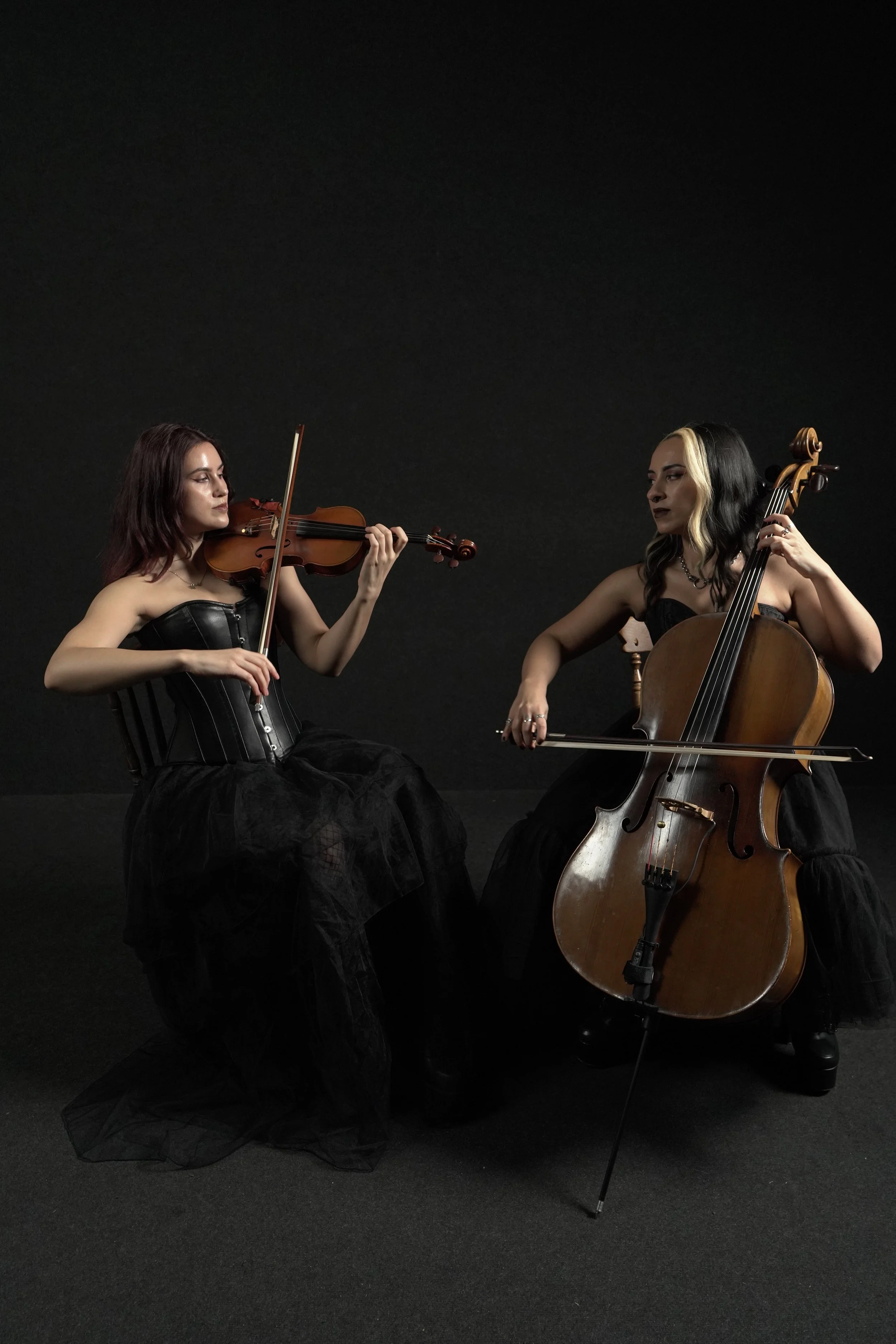 Two women dressed in black gothic style outfits playing violin and cello in a dark studio setting.