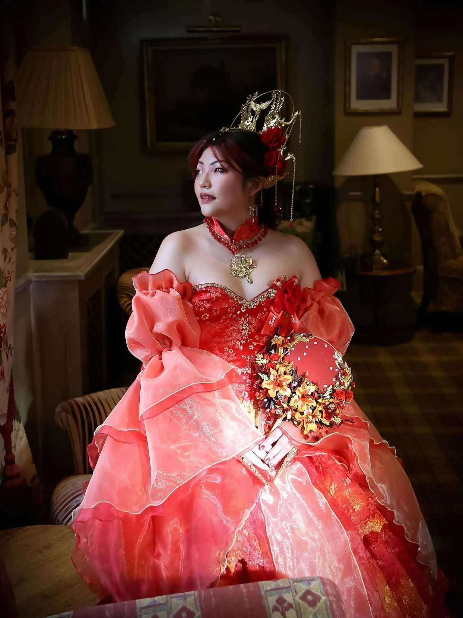 A woman dressed in traditional Asian attire, wearing a red gown with ruffled sleeves, holding a floral red and gold fan, sitting in a warmly lit room with framed pictures and lamps in the background.