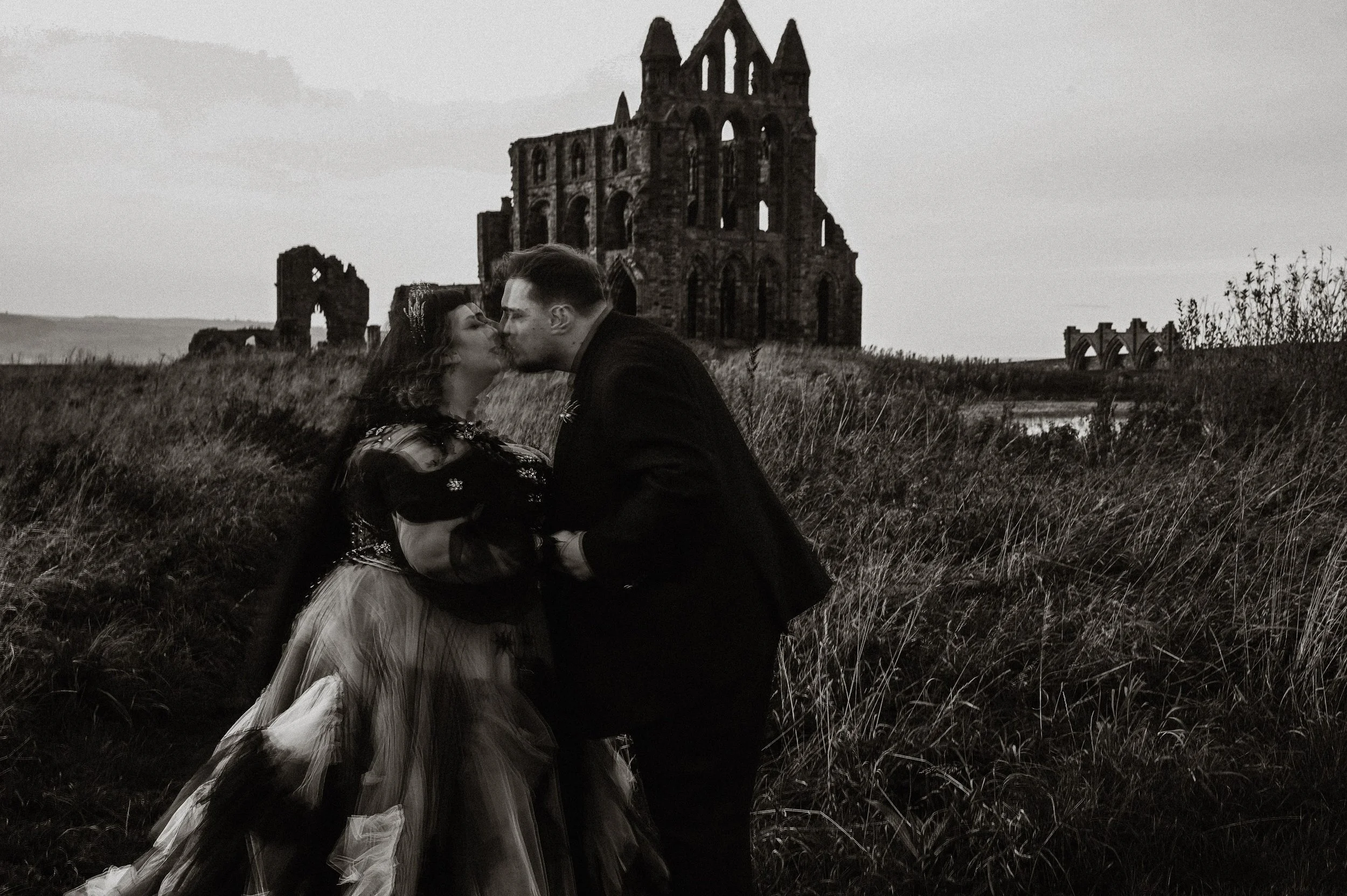 Black and white image. couple share a kiss in the foreground but out of focus and the imposing ruins of Whitby abbey loom in the background