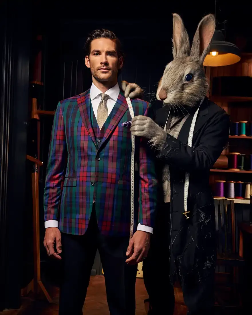 Man in a plaid suit with a white shirt and tie standing next to a rabbit with a human body, dressed in black with a measuring tape, in a tailoring or fashion studio.