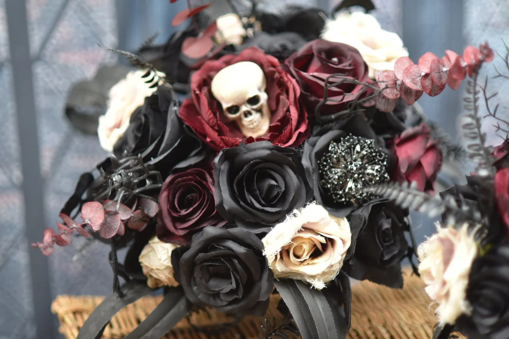 A floral arrangement featuring black, deep red, and cream-colored roses, decorated with a small skull and sparkly embellishments.