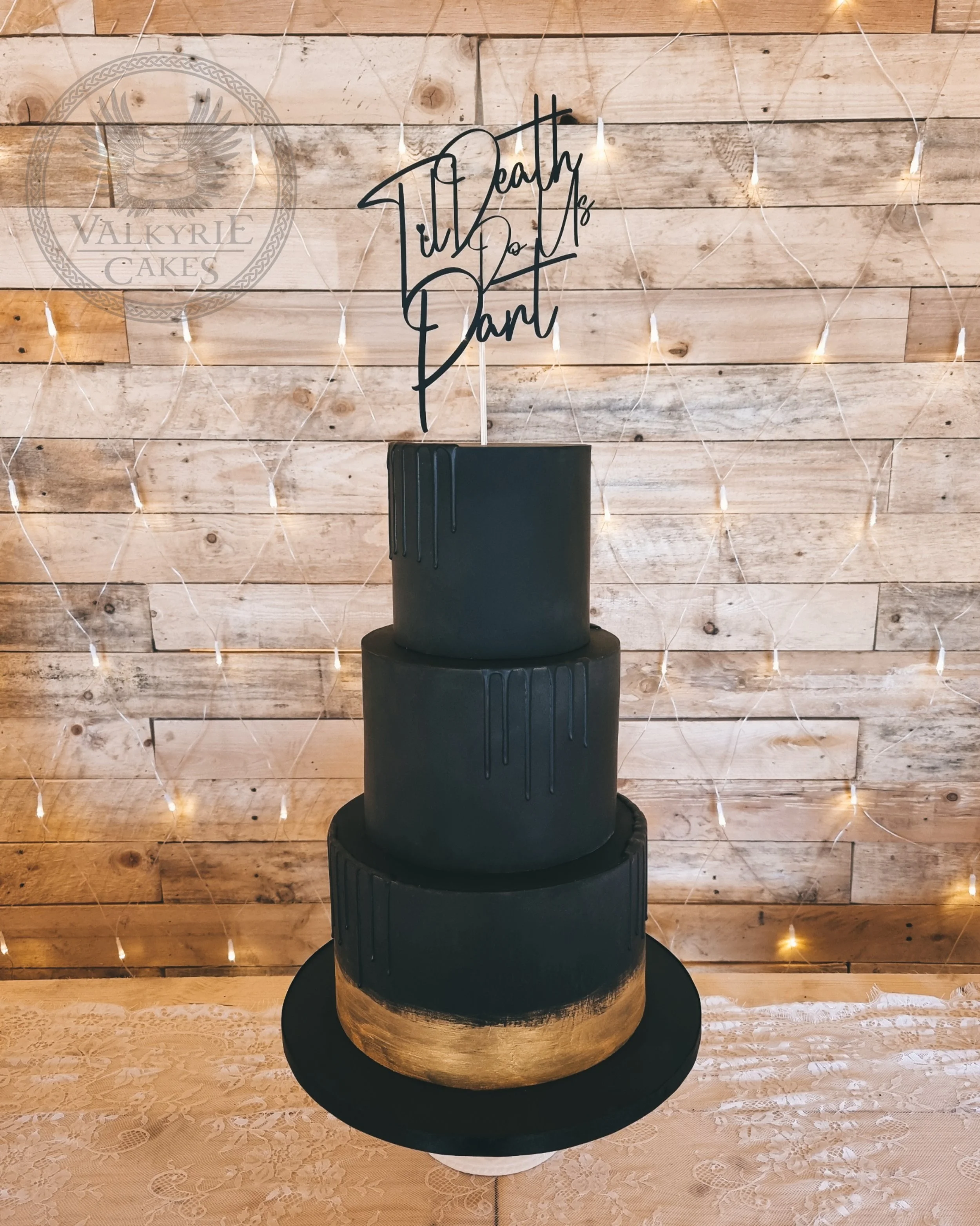 A three-tier black cake with gold accent on the bottom tier, decorated with a black wire cake topper that says 'Still Death Do Us Part,' set against a rustic wood wall with fairy lights.