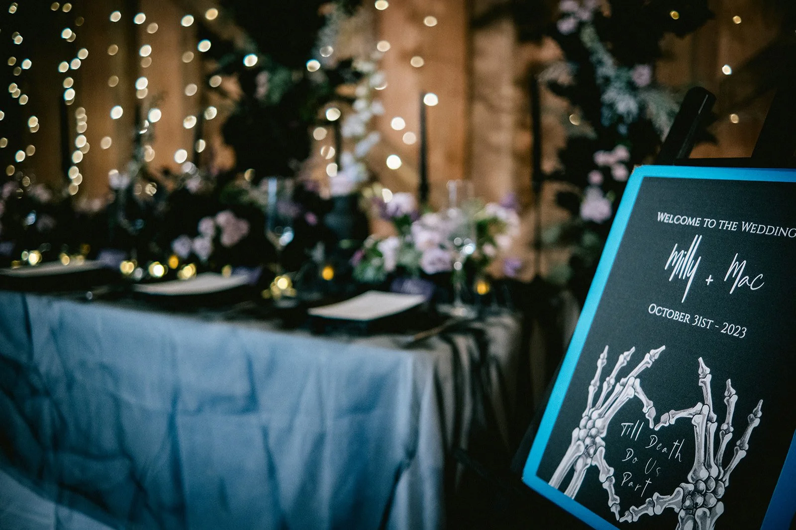 A wedding reception table with floral decorations, fairy lights, and place settings. A sign reads 'Welcome to the wedding Willy + Mac October 31st, 2023 Till Death Do Us Part' with a skeletal hand illustration.