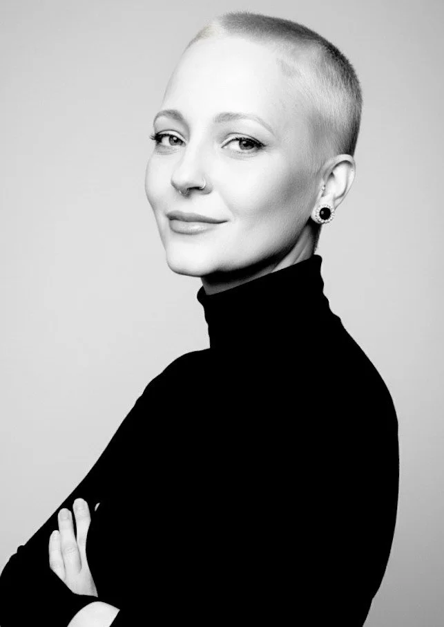 Black and white portrait of a woman with a shaved head, wearing a black turtleneck, with a nose ring and earrings, looking at the camera with a slight smile.