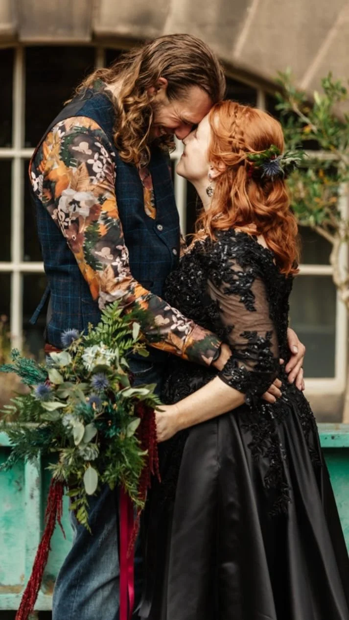 A man and woman share a romantic moment, touching foreheads and smiling at each other in an indoor setting with large windows and greenery. The man has long, wavy hair and wears a floral shirt with a plaid vest, while the woman has red hair styled wi