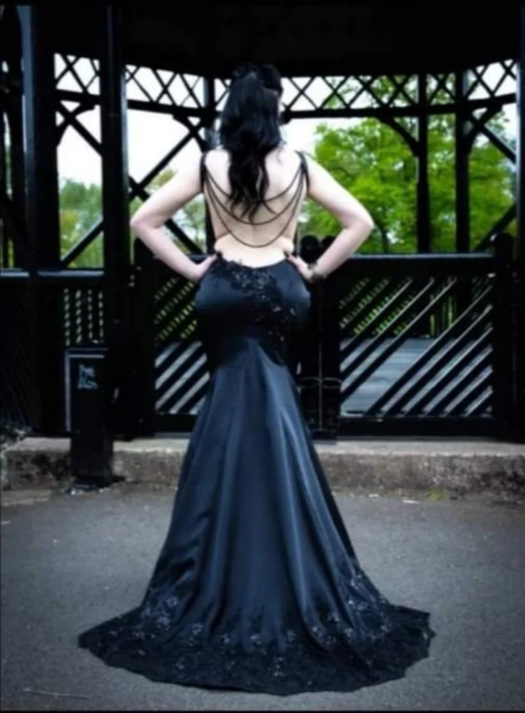 A woman in a backless, black evening gown with lace detailing, standing with her hands on her hips, outdoors near a wooden fence or gate.