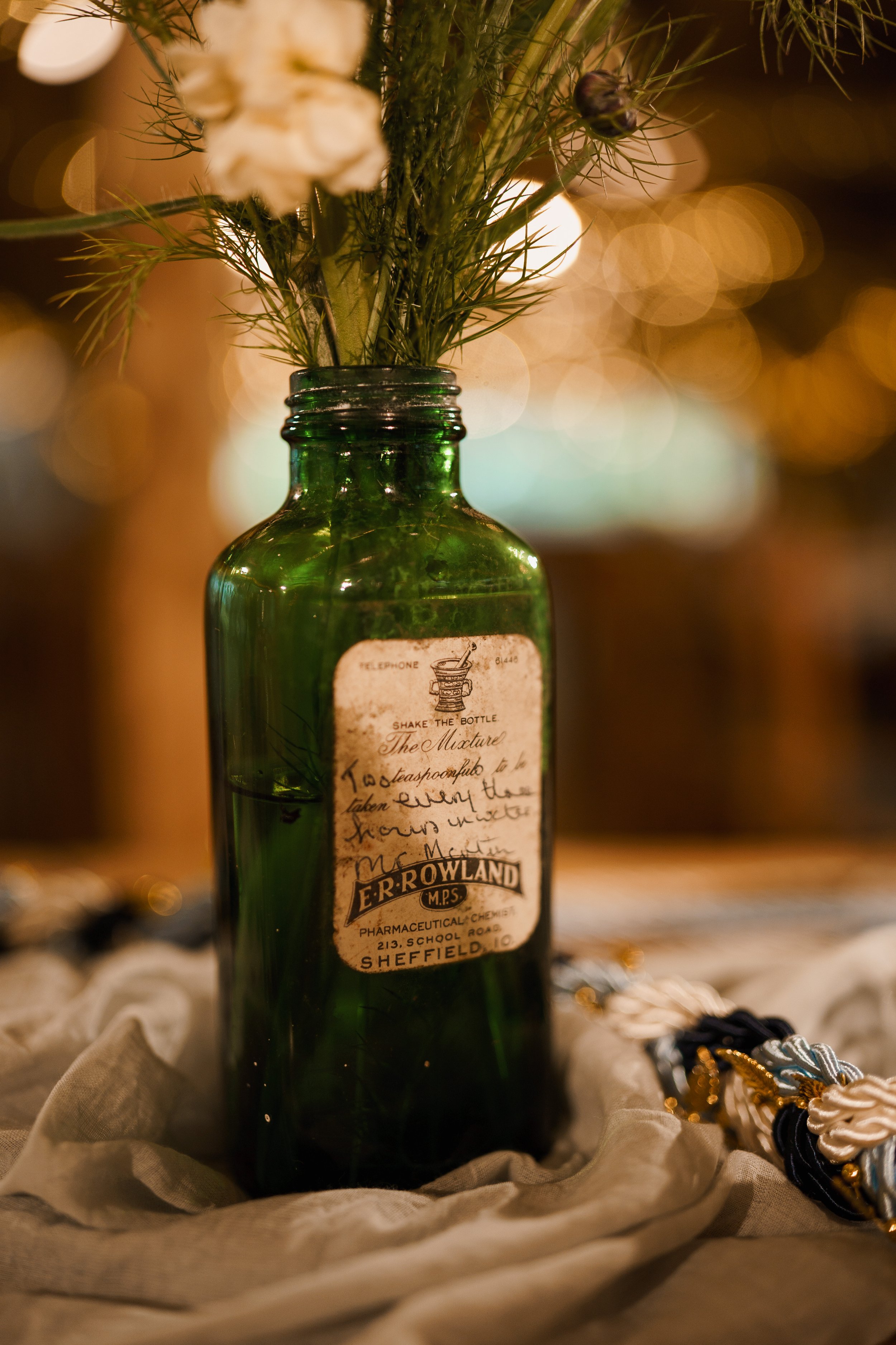 A vintage green glass apothecary bottle with a design and handwritten label, used as a flower vase with white flowers and greenery inside. The bottle is on a beige cloth, with jewelry nearby, against a blurred warm-toned background.