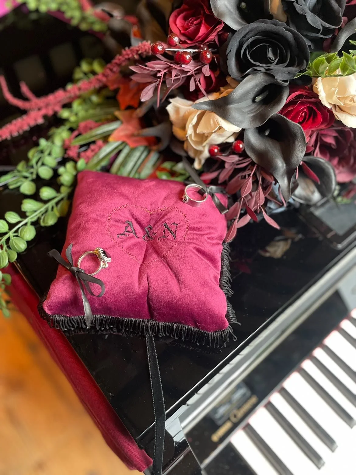 A pink velvet ring pillow with black fringe detailing and tied with black ribbons, placed on a piano with a bouquet of red, black, and cream-colored flowers nearby.