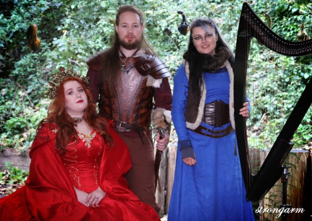 Three people dressed in medieval fantasy costumes outdoors with greenery. A woman in red with a crown and long red hair sits, a man in brown armor with long hair stands, and a woman in a blue dress with black hair and a fur-lined vest stands near a h