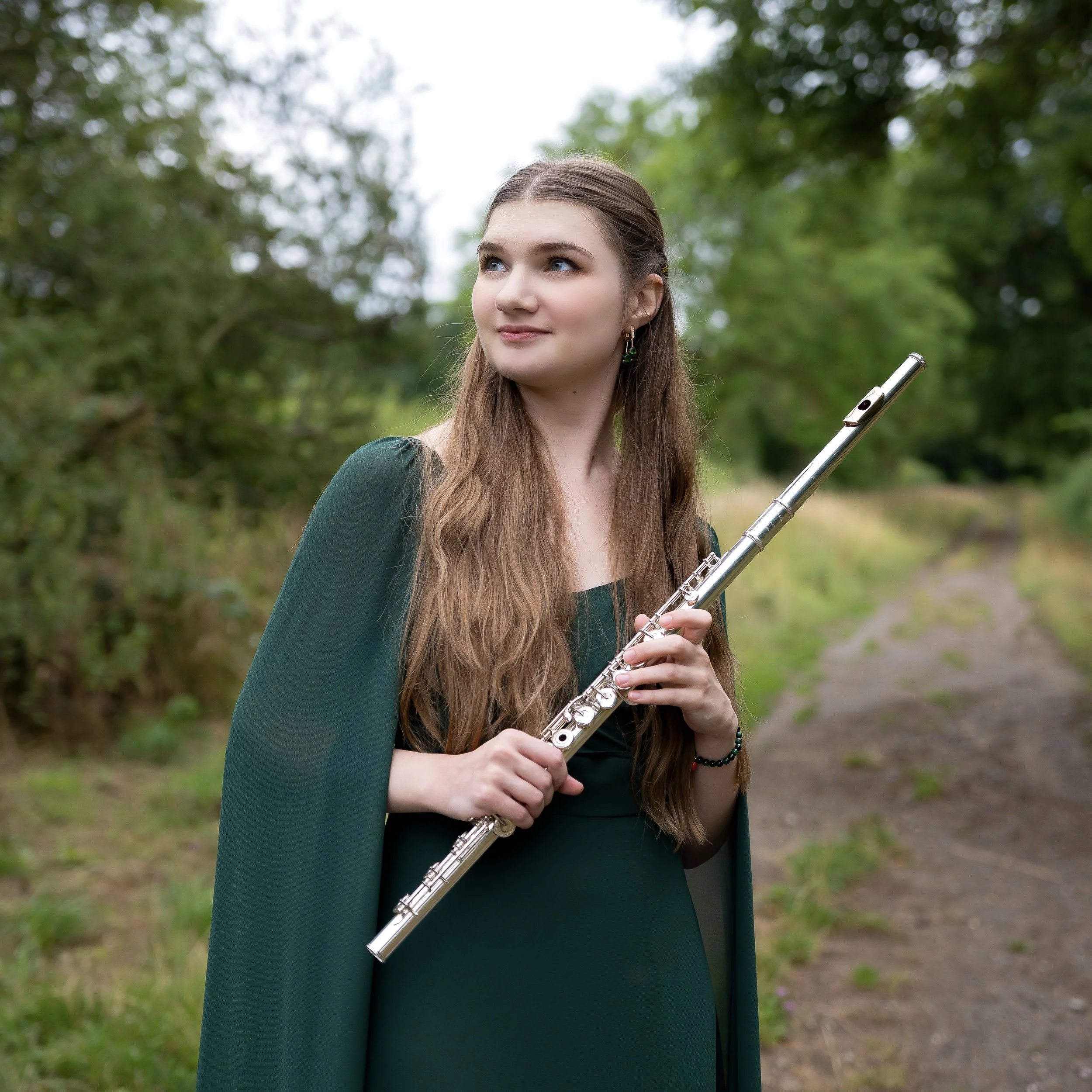 Young woman with long brown hair, wearing a dark green dress and cape, holding a silver flute, standing outdoors on a dirt path surrounded by trees and greenery.