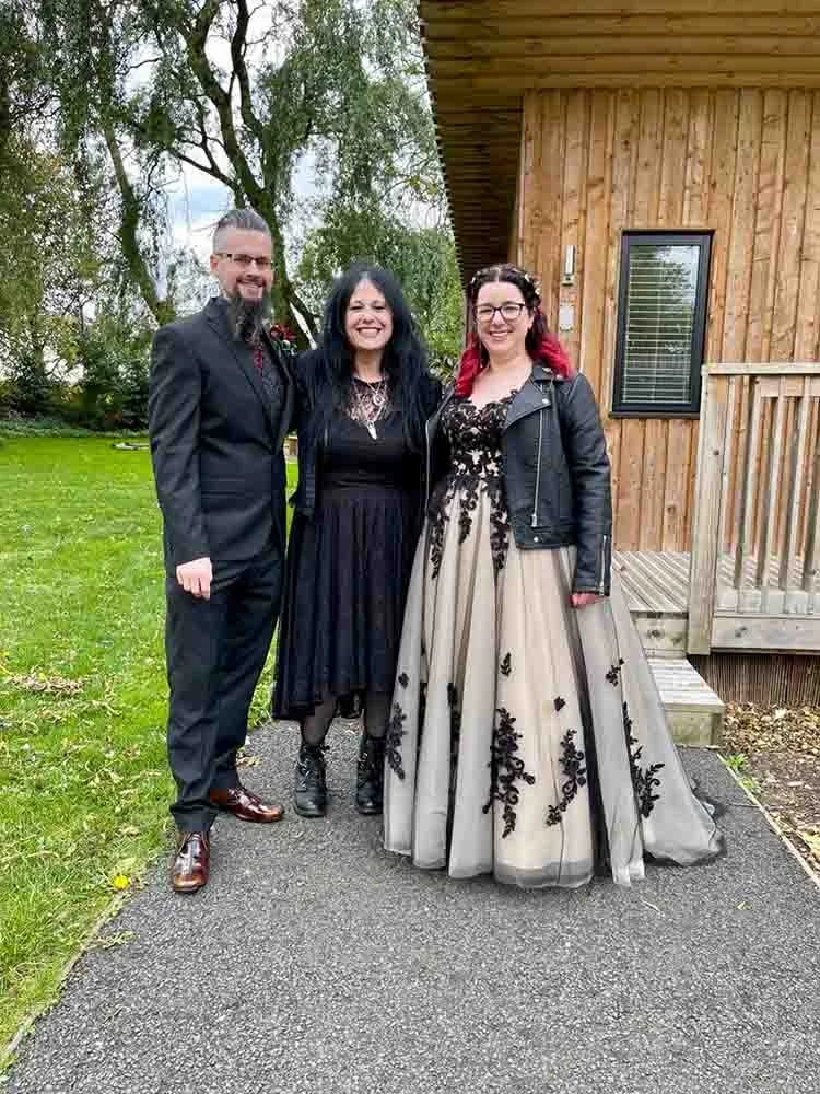 Three people standing outdoors on a path in front of a wooden house, dressed in formal attire. The man on the left is wearing a dark suit, the woman in the middle is in a black dress with a necklace, and the woman on the right is wearing a beige gown