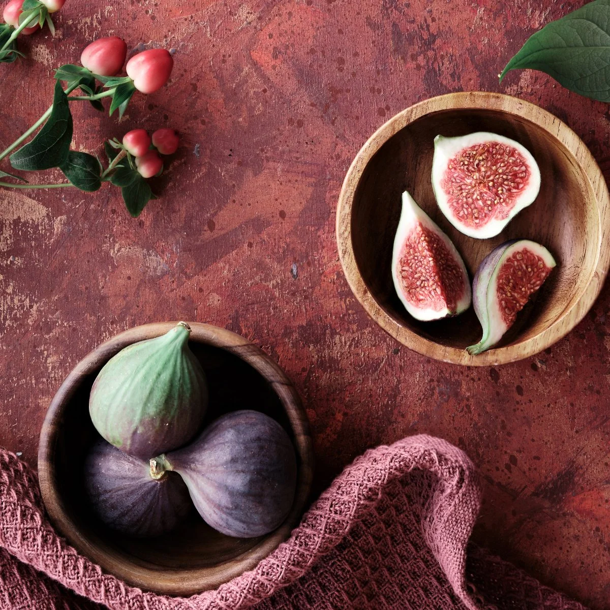 A rustic red surface with a bowl of three figs, a wooden bowl with halved figs showing red interior, and a sprig of pink berries with green leaves.