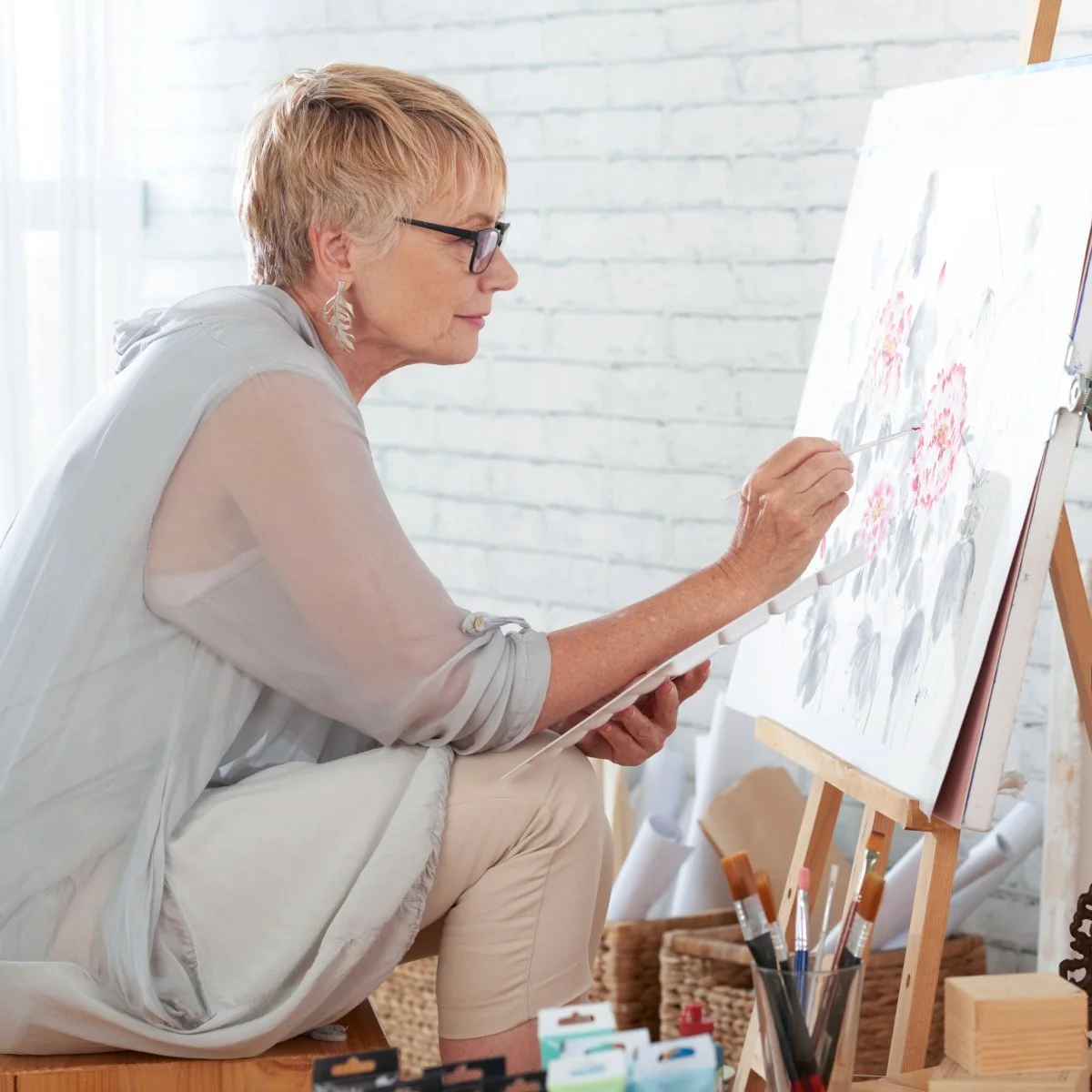 An older woman with short reddish hair, glasses, and earrings, sitting on a wooden stool, painting pink flowers on a canvas in a bright, white room.