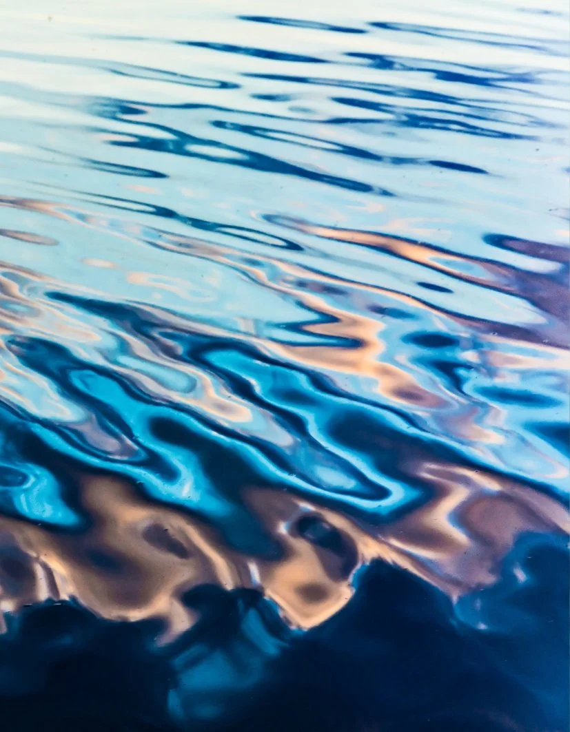 Close-up view of water surface with reflections and ripples, displaying shades of blue, beige, and pink.
