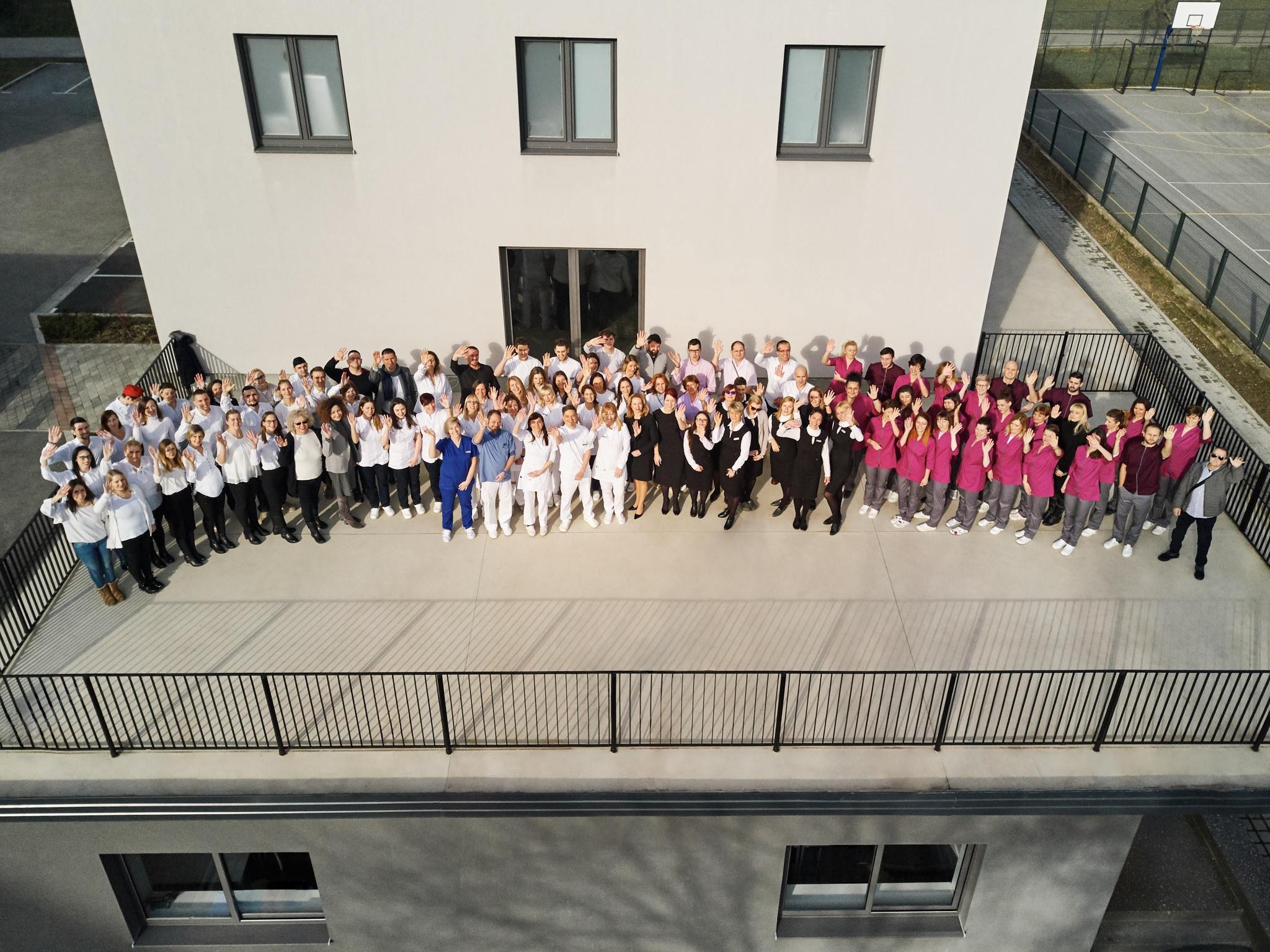 A large group of healthcare professionals, including doctors and nurses, gathers on a rooftop terrace, waving at the camera during daytime. Some are in white coats, others in pink scrubs, and additional staff in casual and formal attire.