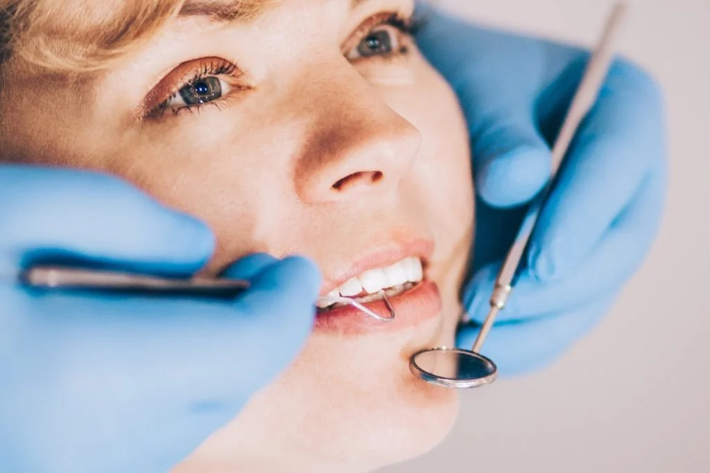 Close-up of a woman at the Dr Wael Fekry Dental Clinic, with a dental mirror in her mouth, as the dentist uses dental tools. The woman is smiling slightly with her teeth visible and appears to be relaxed.